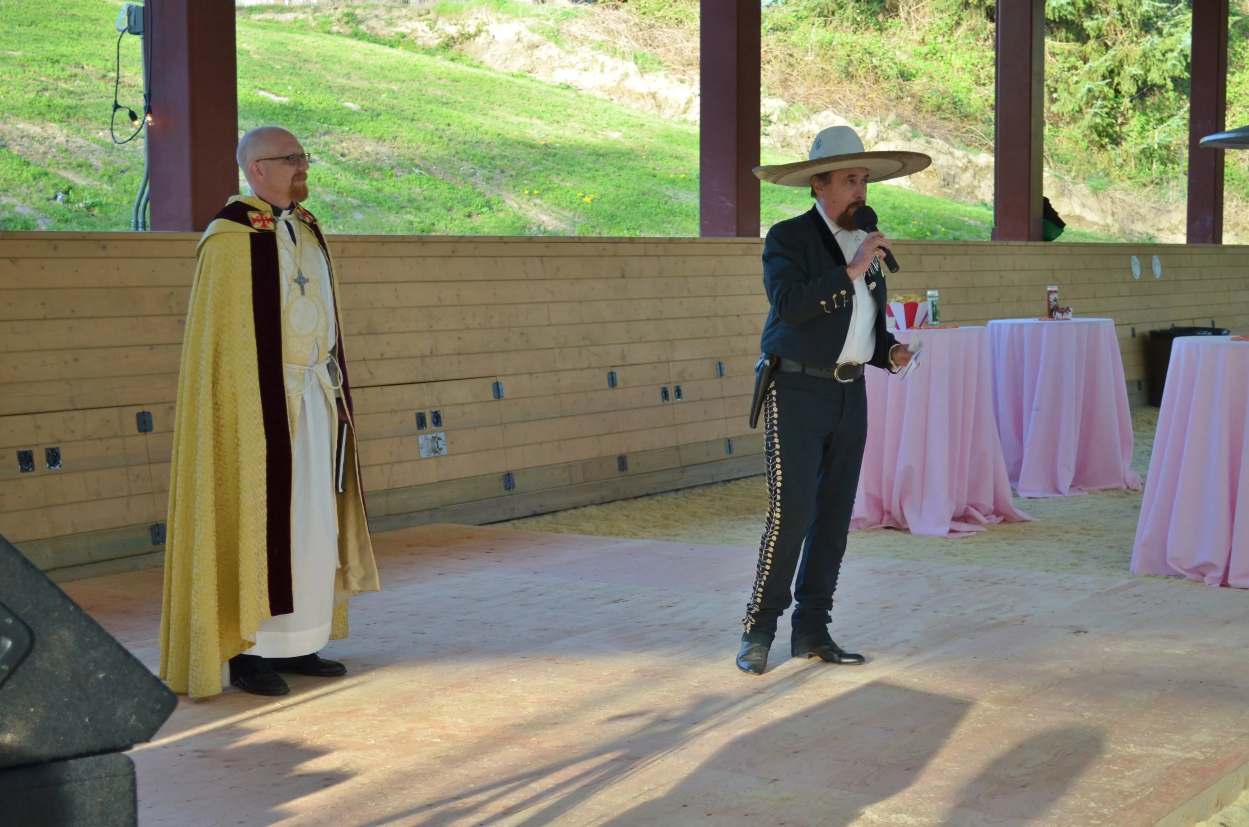 A man dressed as a mariachi in a traditional sombrero, black suit, and decorative pants holding a microphone, speaking to an audience. A priest or clergy member in religious robes and a cape stands nearby, observing. The setting appears to be an outd