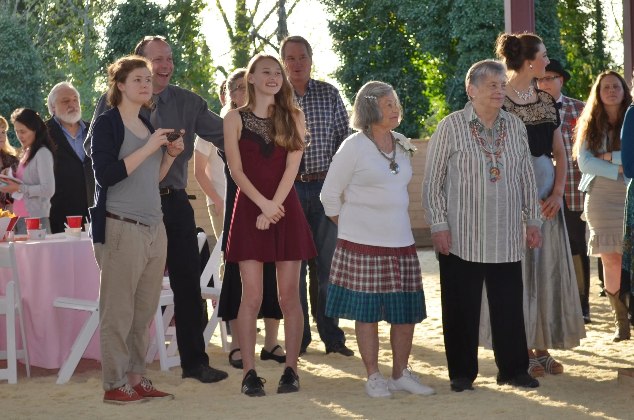 Group of people, including children and older adults, gathered outdoors at a social event, standing on sand with trees in the background, some smiling and others looking around.
