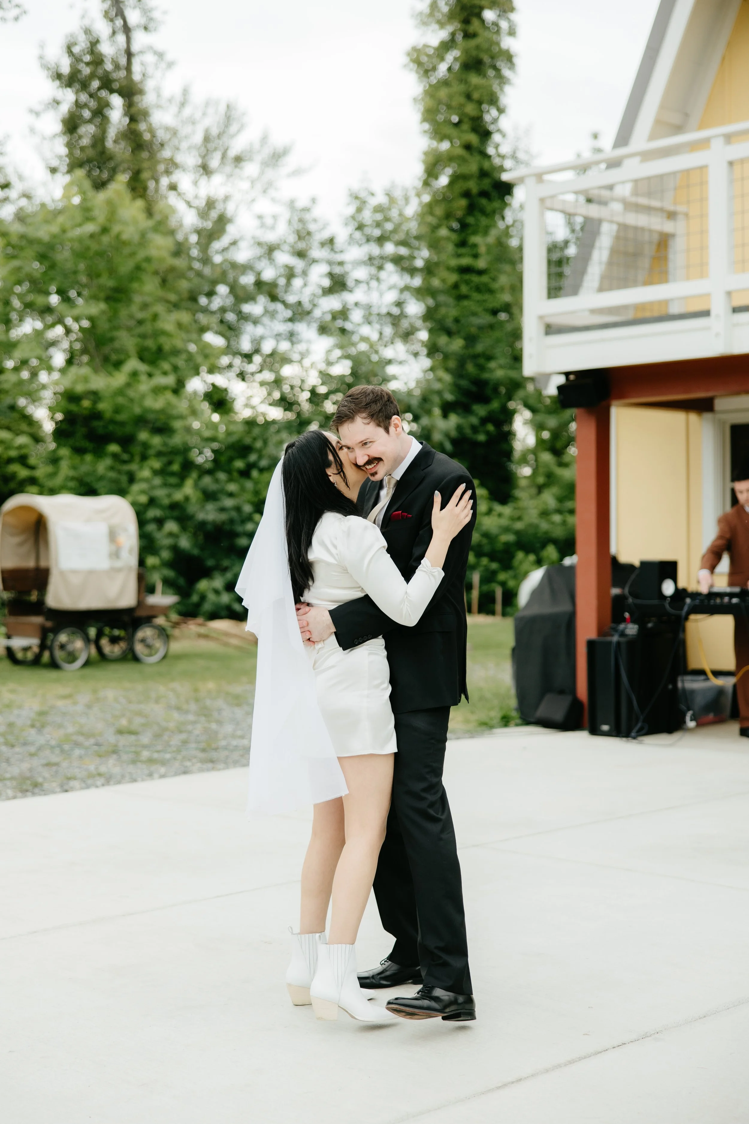 A bride and groom hug and smile at each other outdoors during their wedding, with a DJ and a yellow house in the background.
