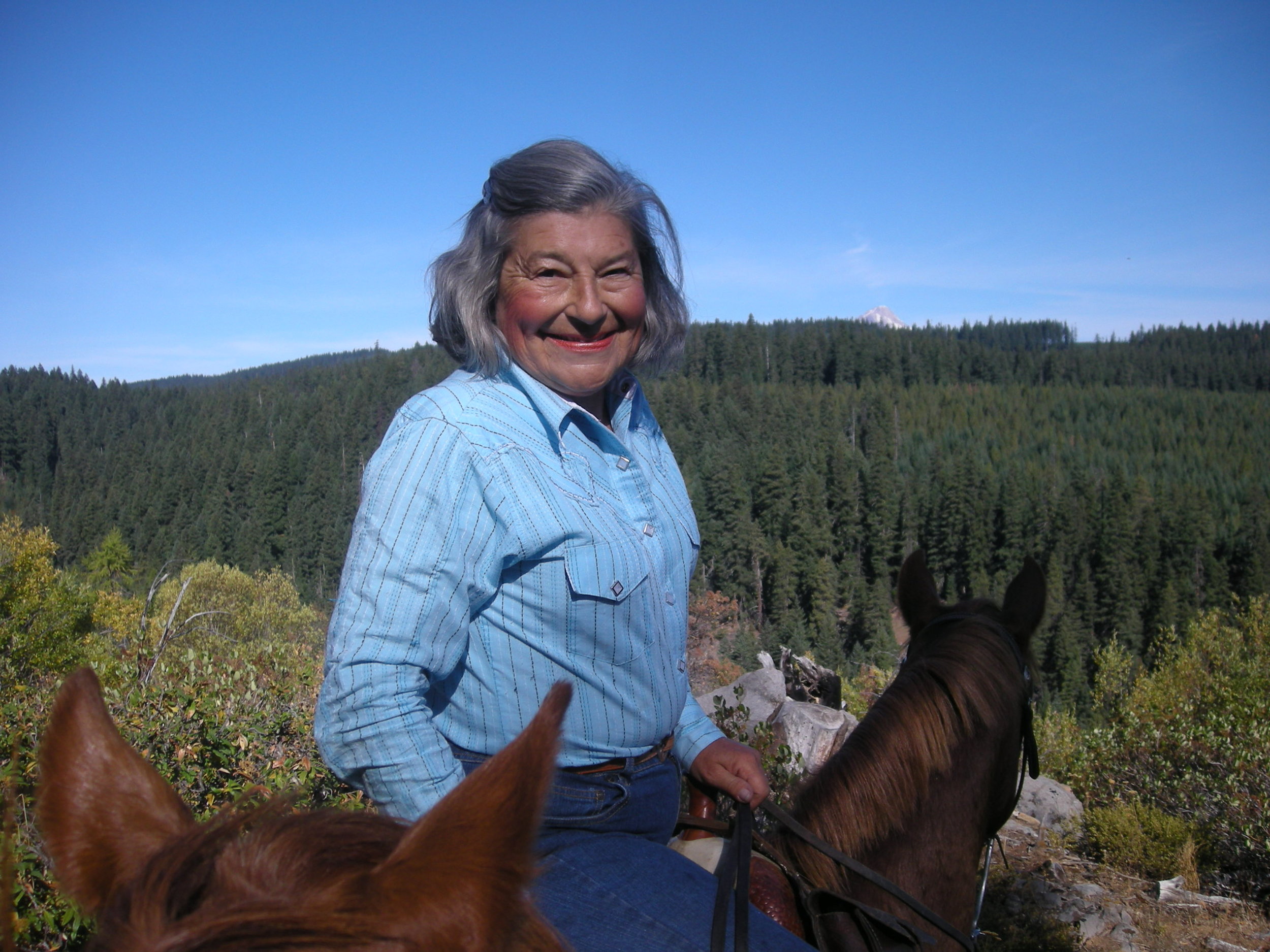 An elderly woman riding a horse outdoors in a forested area with a mountain in the background, smiling at the camera.