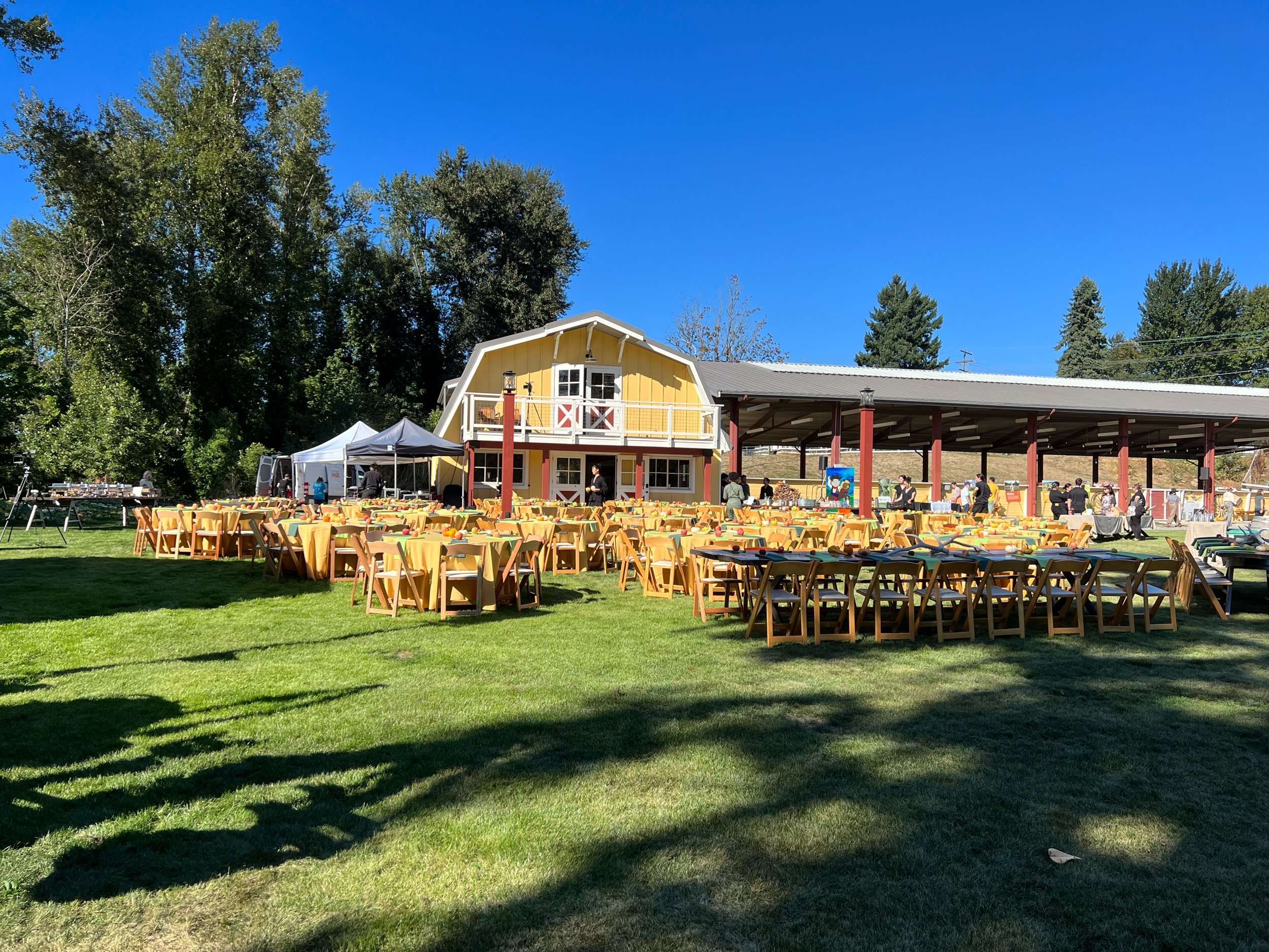 Outdoor event setup with round tables and chairs on a grassy area, yellow barn building, tents, and people preparing for a gathering, under a clear blue sky.