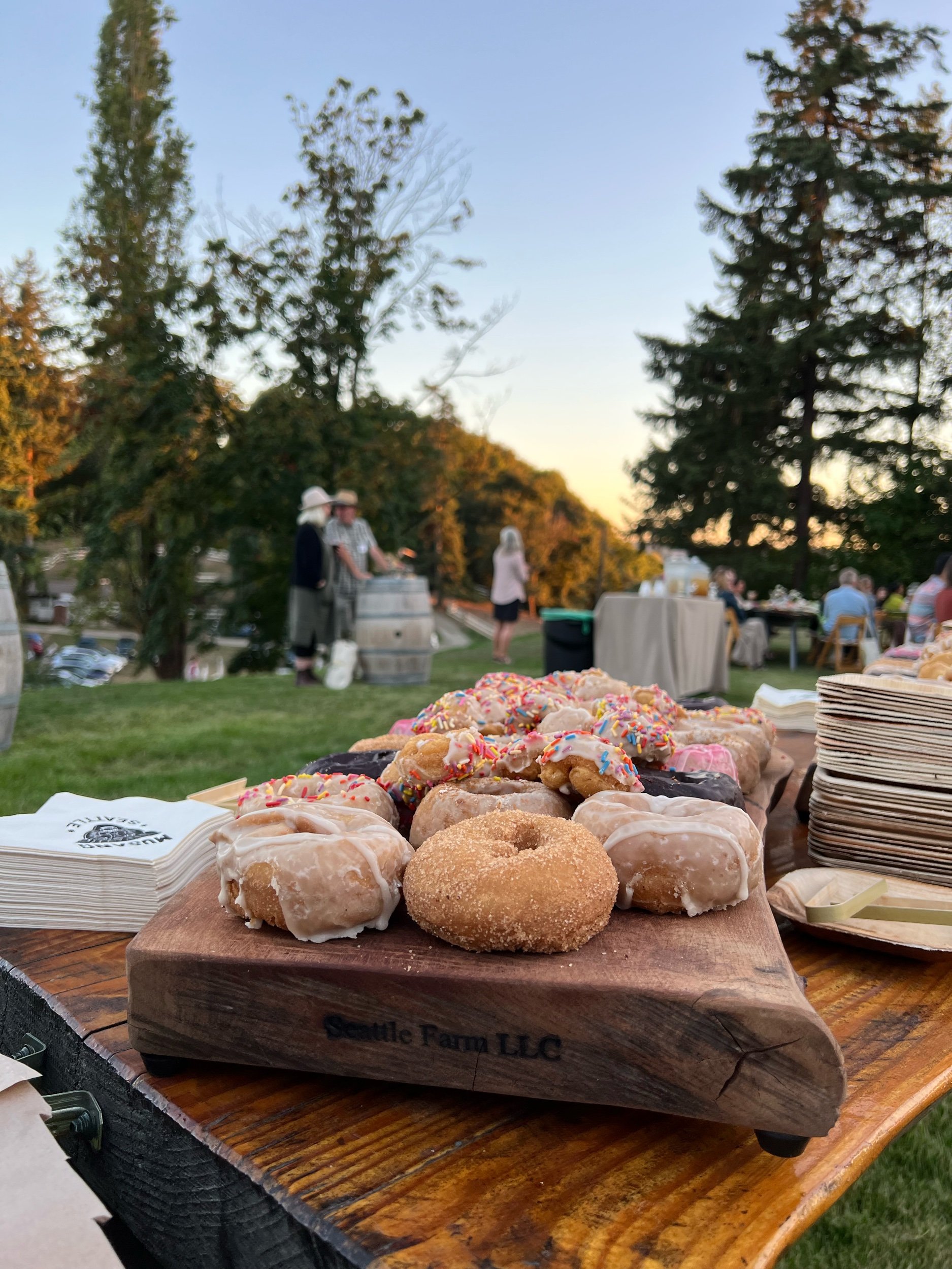 Assorted glazed and frosted donuts with sprinkles on a wooden serving board at an outdoor gathering, with people socializing in the background during sunset.