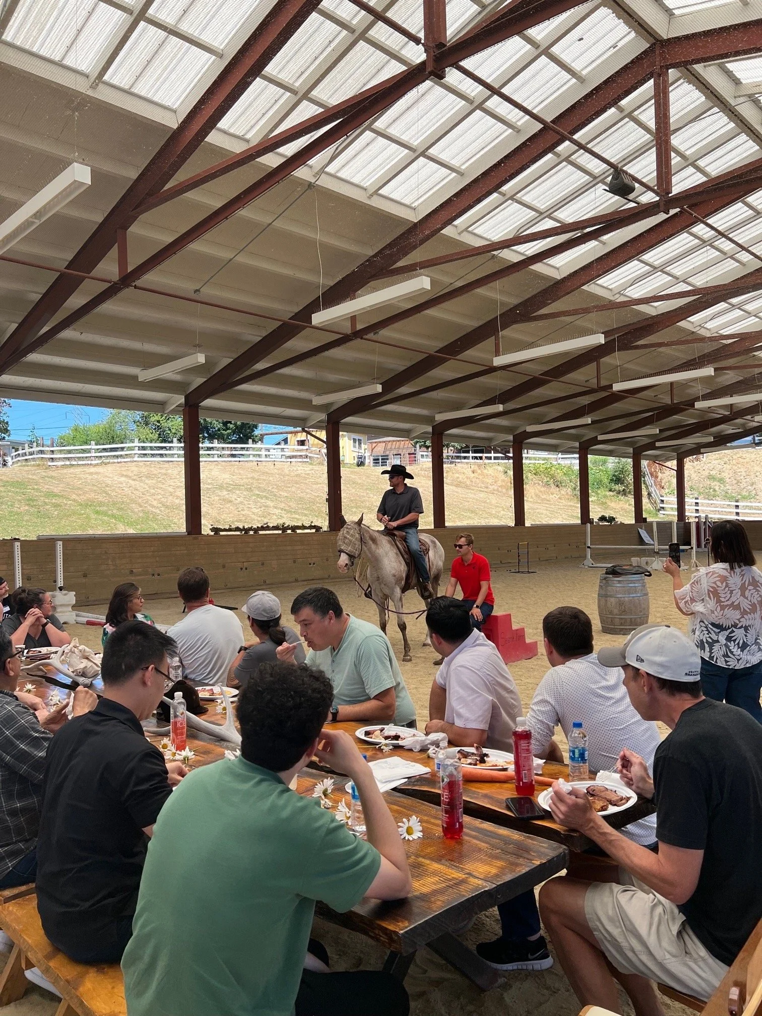 People sitting at a long table under a covered pavilion watching a horseback riding demonstration, with a person on horseback and another person guiding the horse.