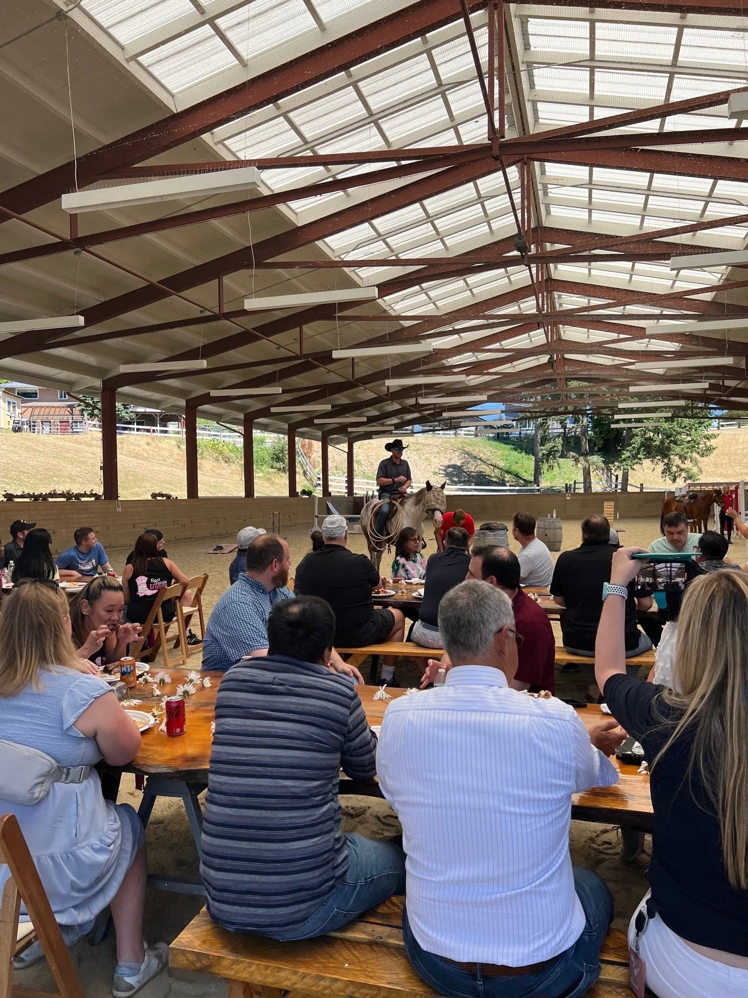 People sitting at picnic tables in an outdoor covered arena, watching a person on horseback under a roof supported by red beams.