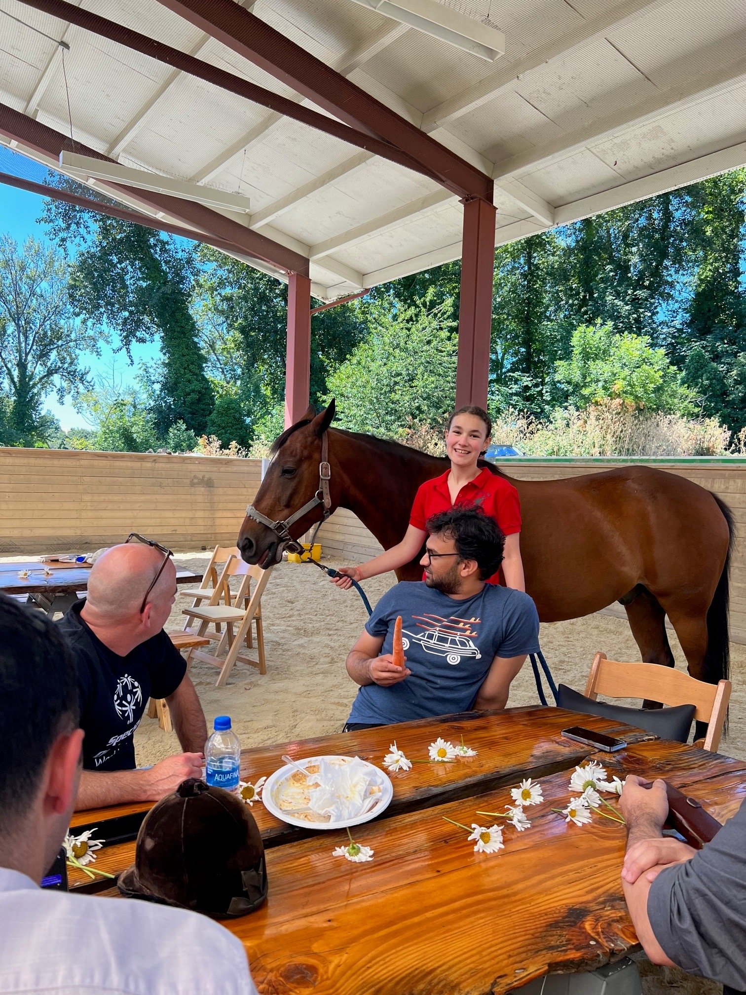 A group of people sitting at a wooden picnic table with white daisies on it, outside a semi-covered area, with a young woman in a red shirt standing beside a brown horse, holding its reins, and conversing with the people at the table. There are trees