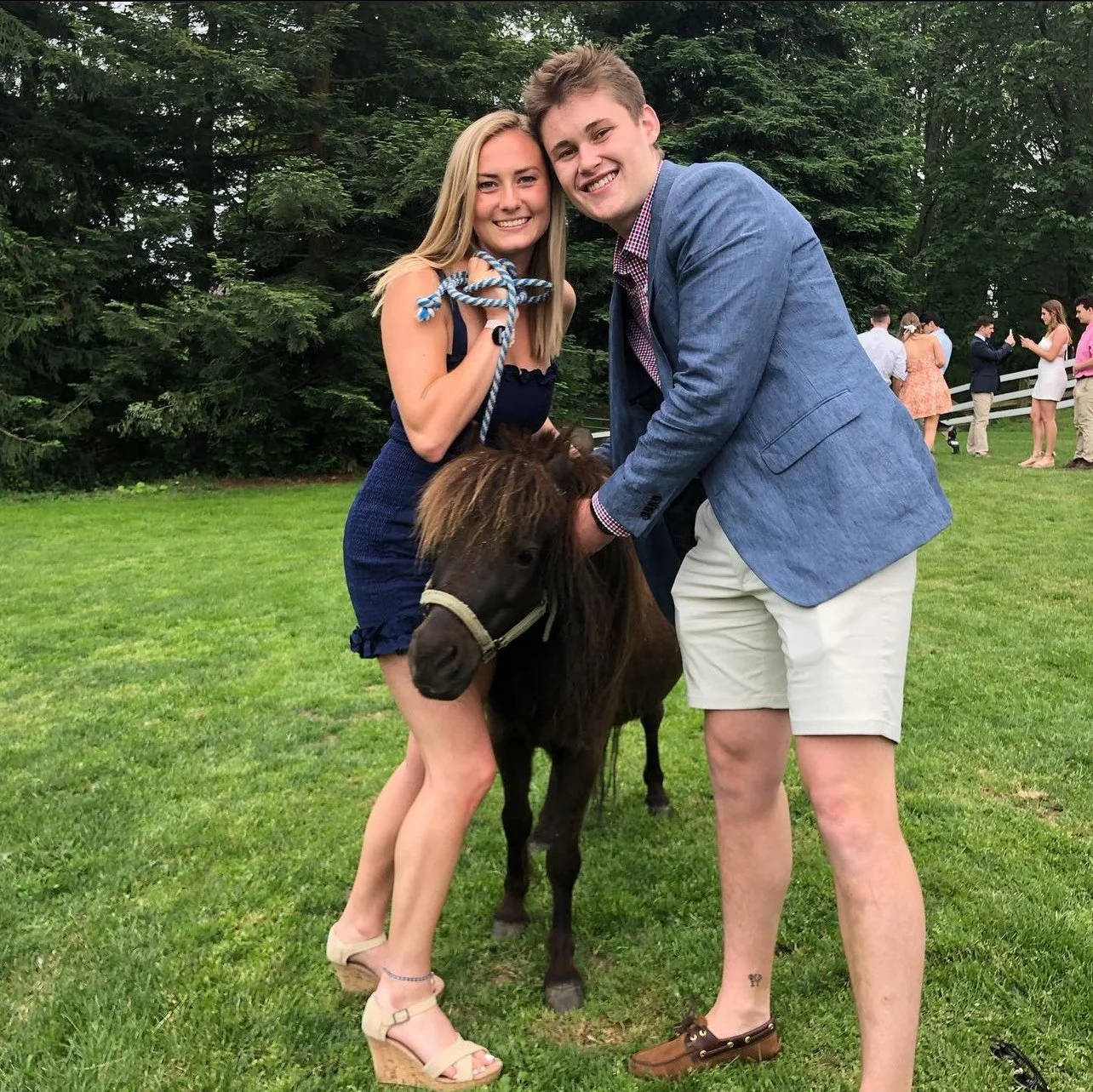 Two smiling young adults, a woman in a dark blue dress and a man in a blue blazer and light shorts, holding a small pony on a grassy area with trees in the background, and several people standing in the distance.