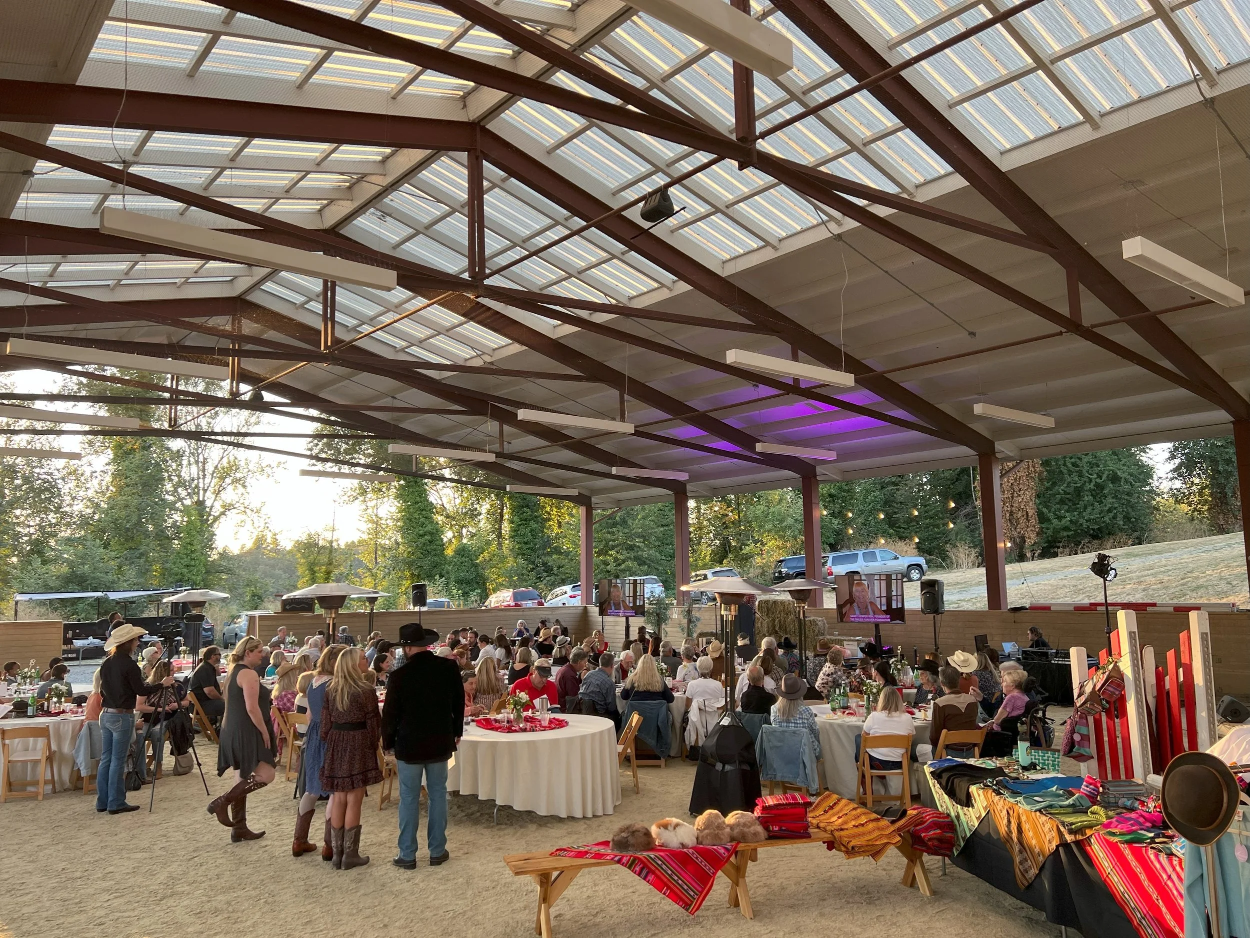An indoor event with many attendees seated at round tables with white tablecloths, and some standing around. The venue has a high, open ceiling with exposed beams and large windows or openings. There are outdoor elements visible through the openings,