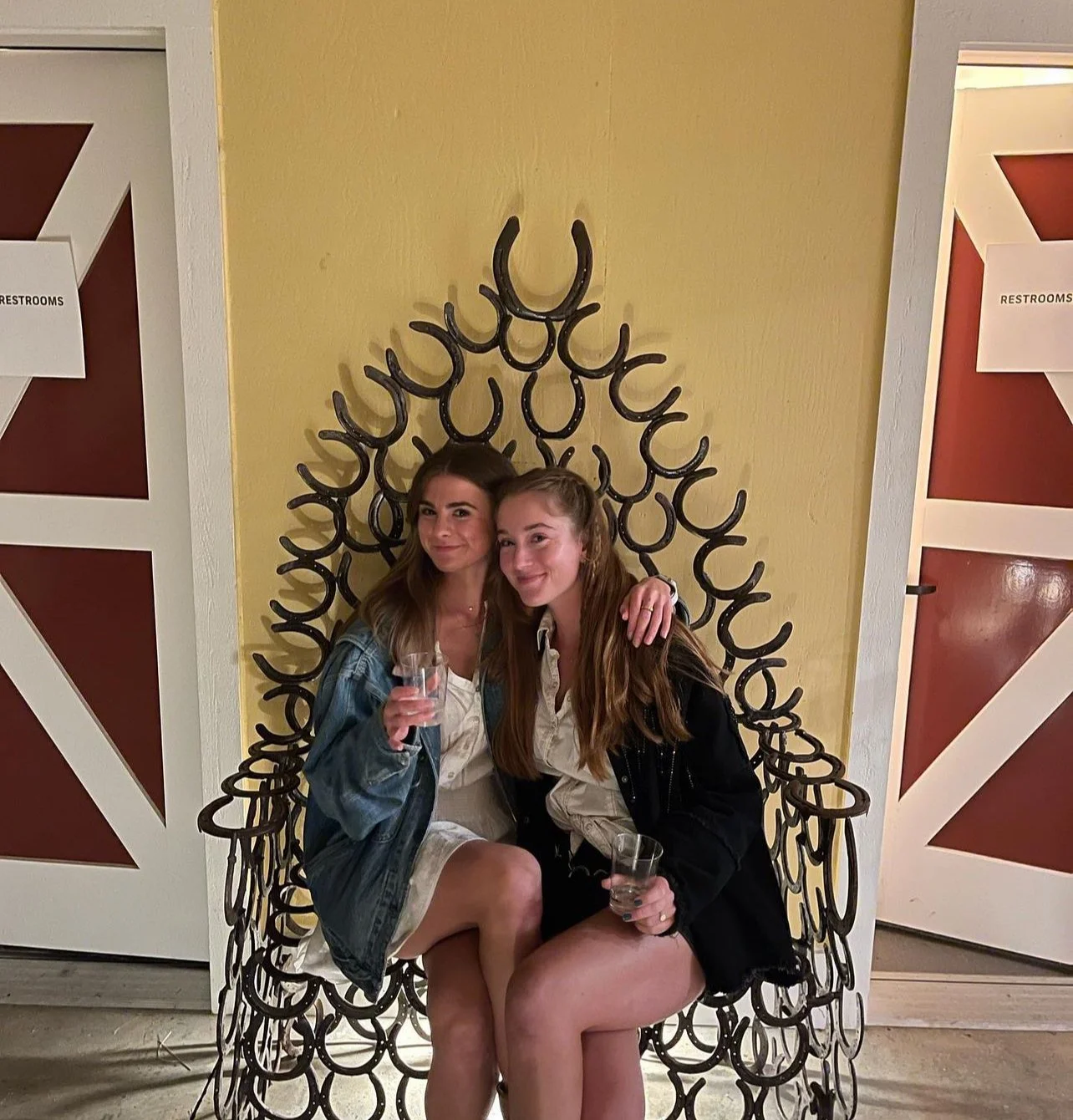 Two young women sitting on a decorative metal chair, smiling, holding clear glasses, with a yellow wall and bathroom signs in the background.