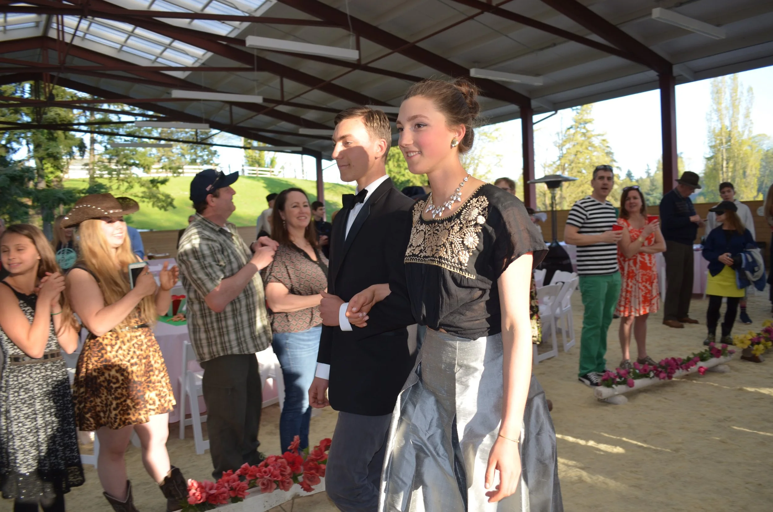 A young man in a tuxedo and a young woman in a black and silver dress are walking together at a social event. People are watching and smiling in the background, with some taking photos. The setting appears to be a covered outdoor space with greenery 
