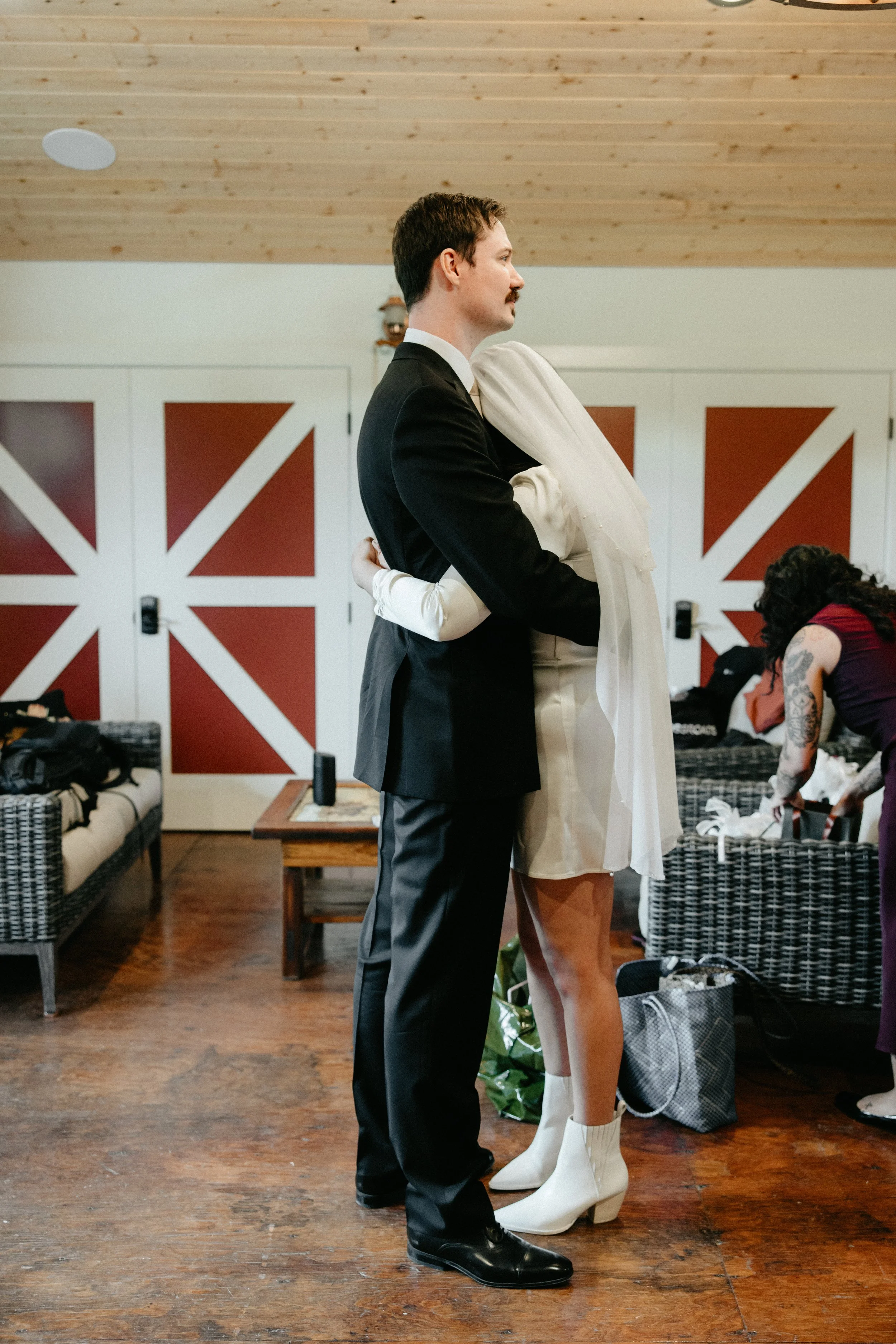 A man in a black tuxedo embracing a woman in a white dress and cowboy boots in a room with wooden floors and red barn doors.