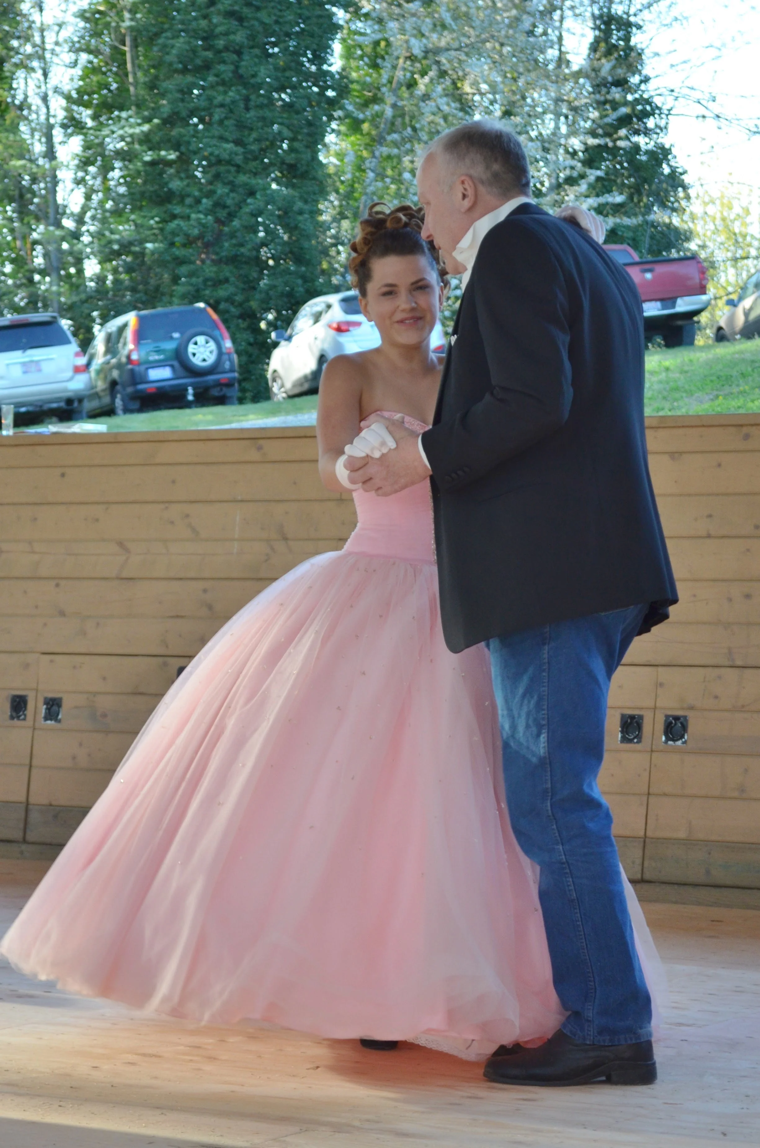 A young woman in a pink ball gown dancing with an older man in a black blazer and blue jeans during an outdoor event.