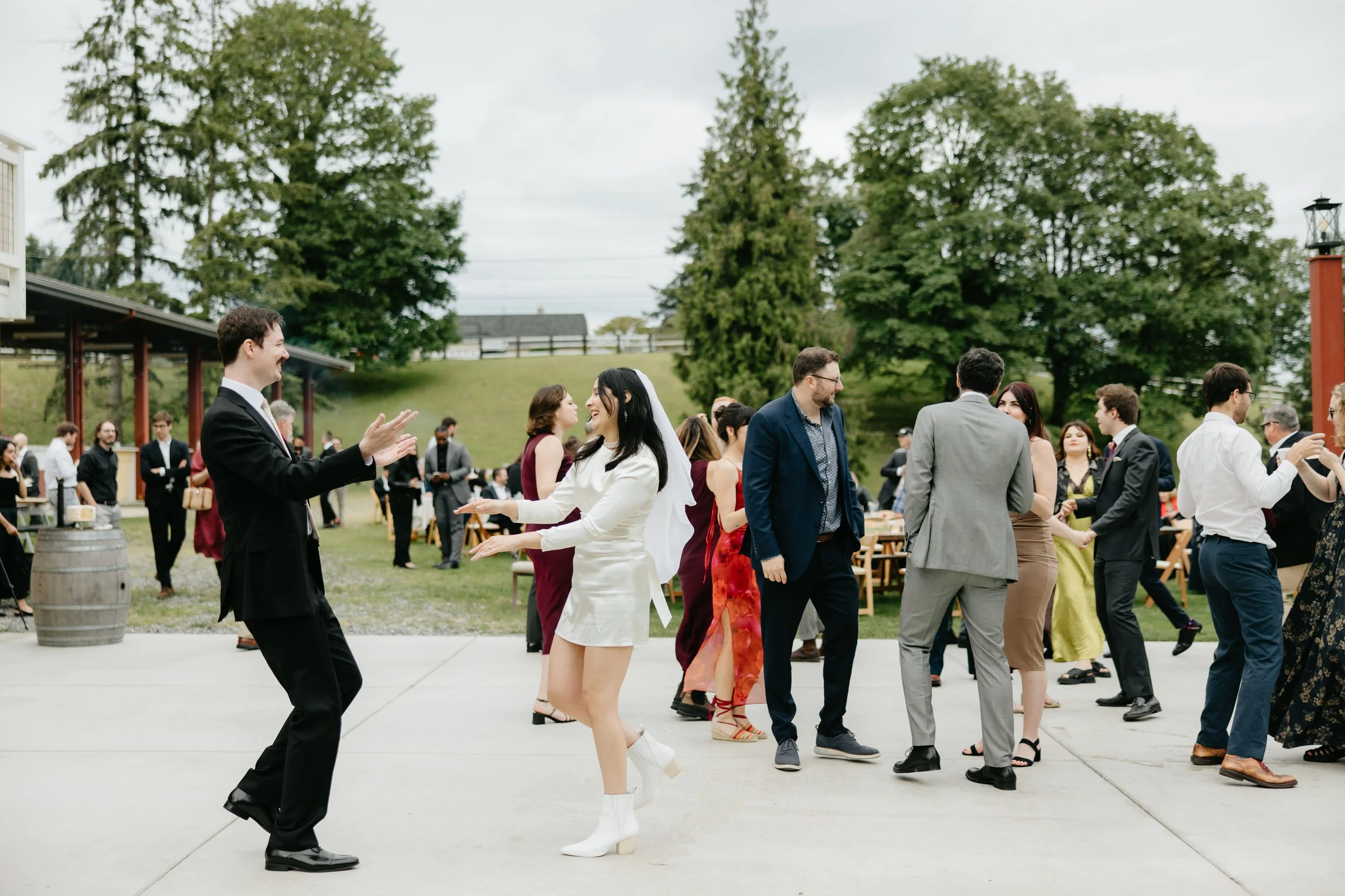People dancing outdoors at a wedding reception, with a green grassy hill and trees in the background.