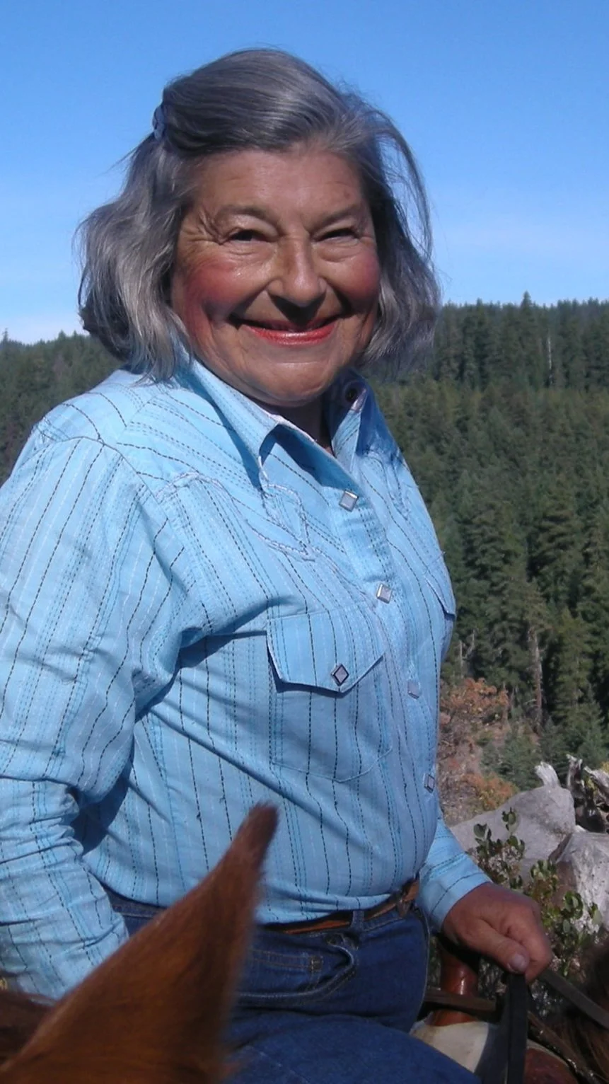 Gloria is smiling outdoors in front of a tree-filled landscape, wearing a blue plaid shirt. in idaho
