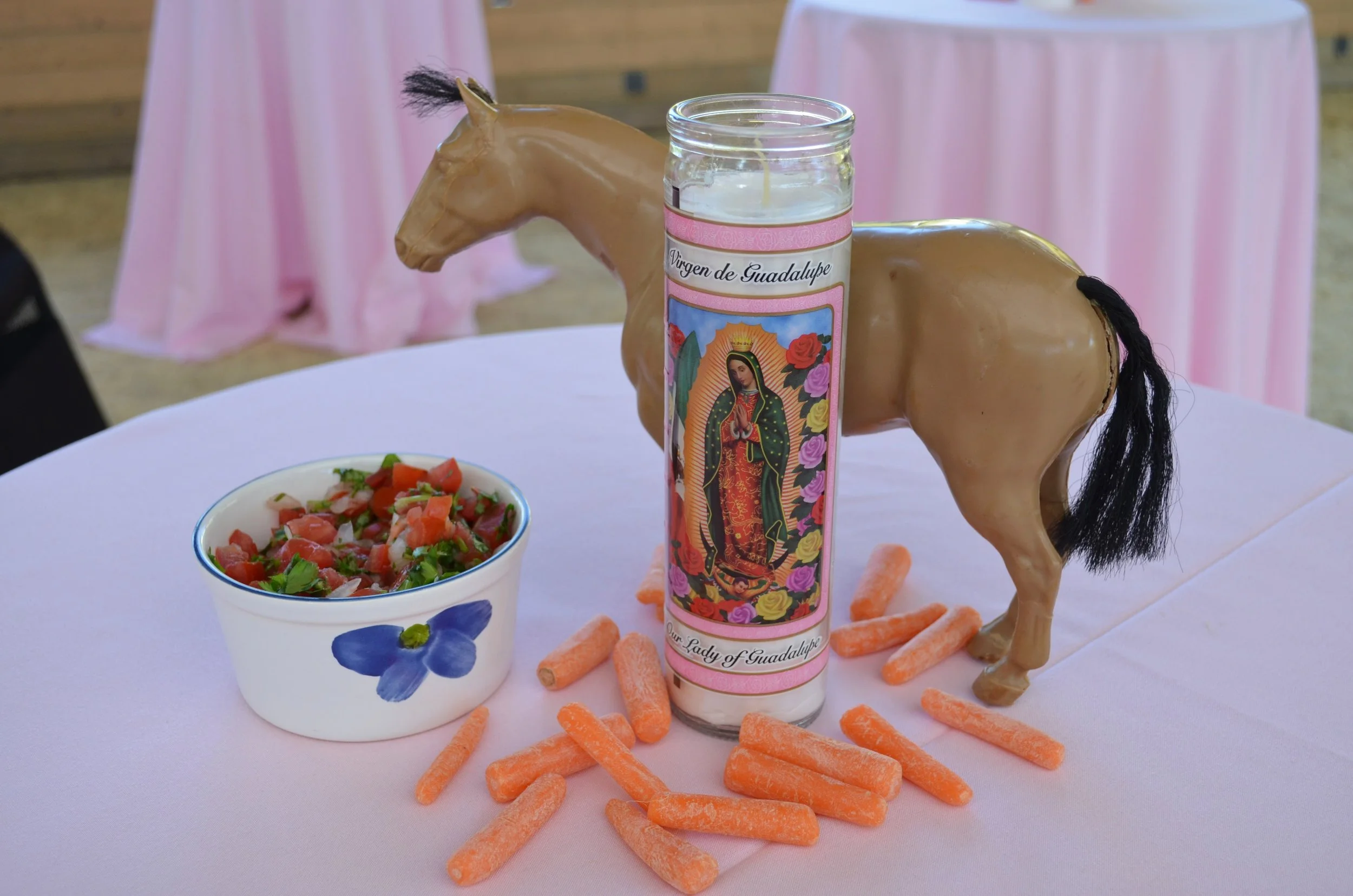 Table display with a plastic horse figurine and a candle decorated with Our Lady of Guadalupe, a bowl of salad with tomatoes and herbs, and scattered baby carrots.