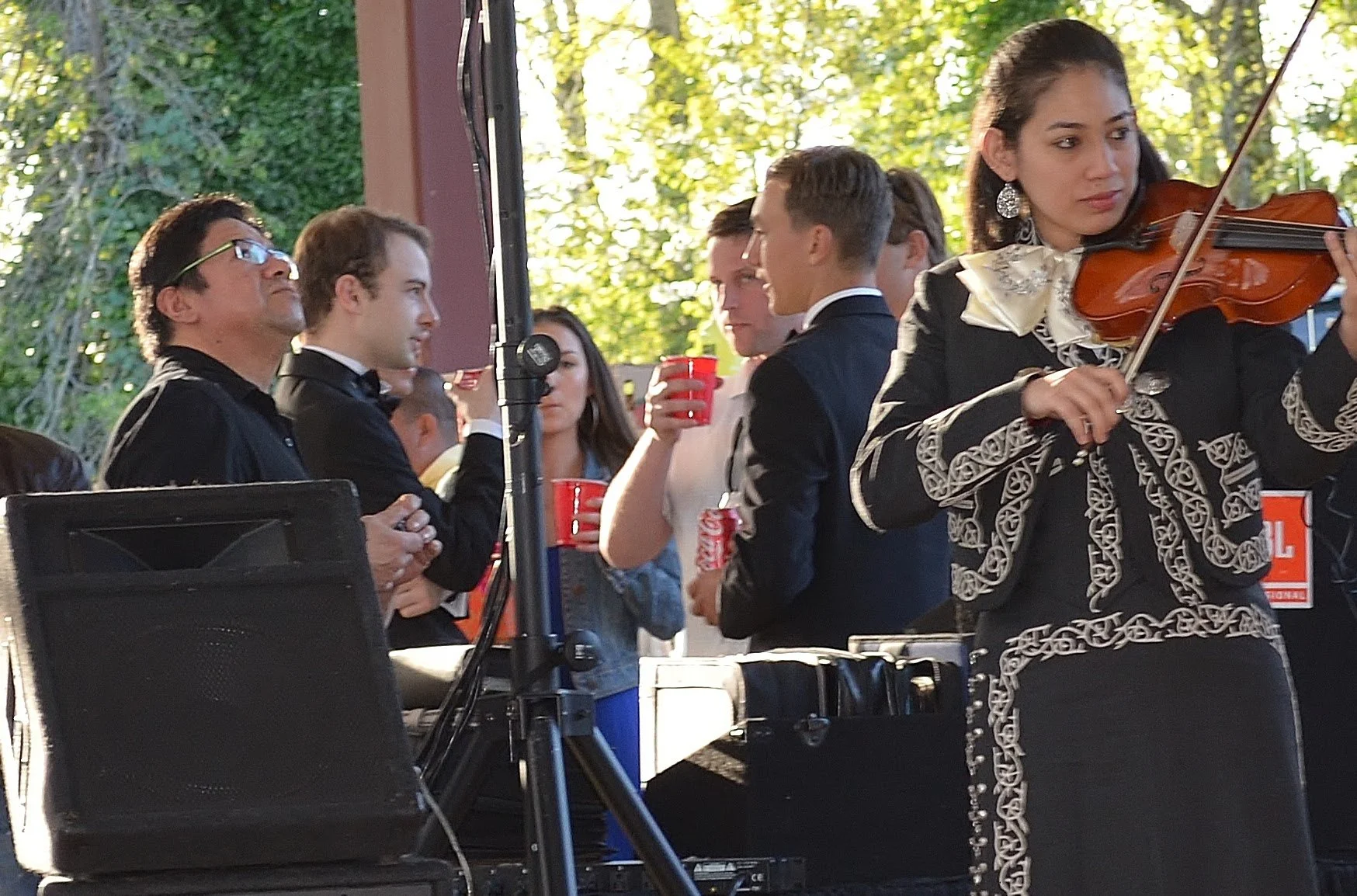 A woman playing the violin at an outdoor concert, with several people in formal attire holding red cups in the background.