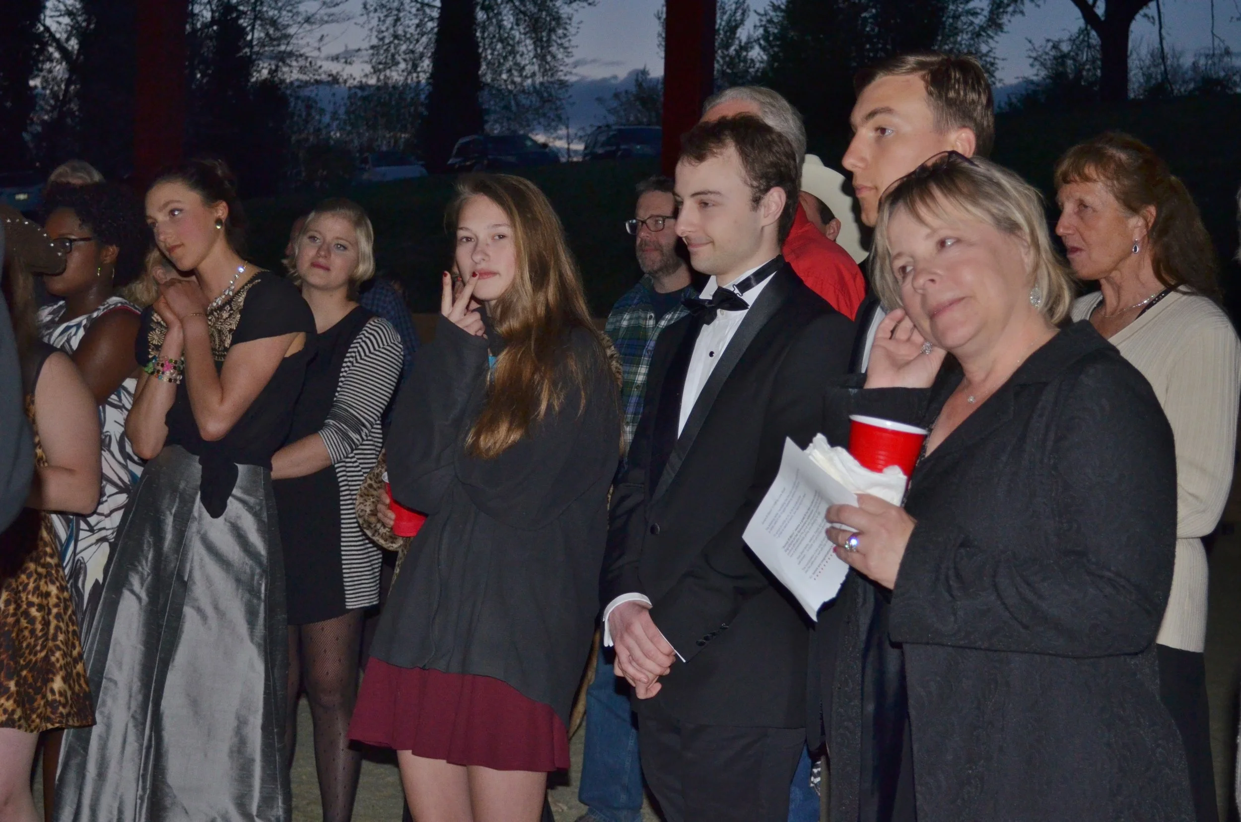 Group of people at an outdoor evening event, some dressed formally, standing in a line and listening attentively.