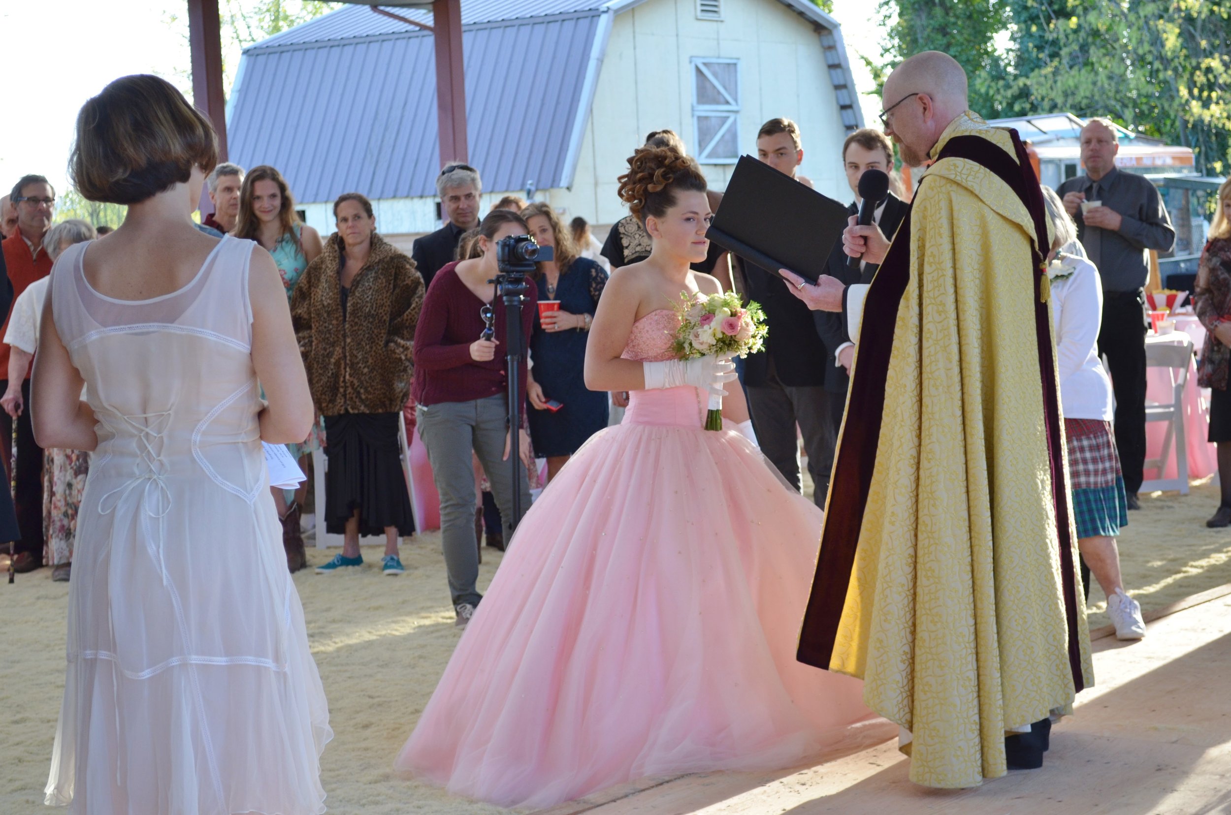 A wedding ceremony outdoors with a bride in a pink ball gown holding a bouquet, standing before a officiant dressed in ceremonial robes, with guests watching.