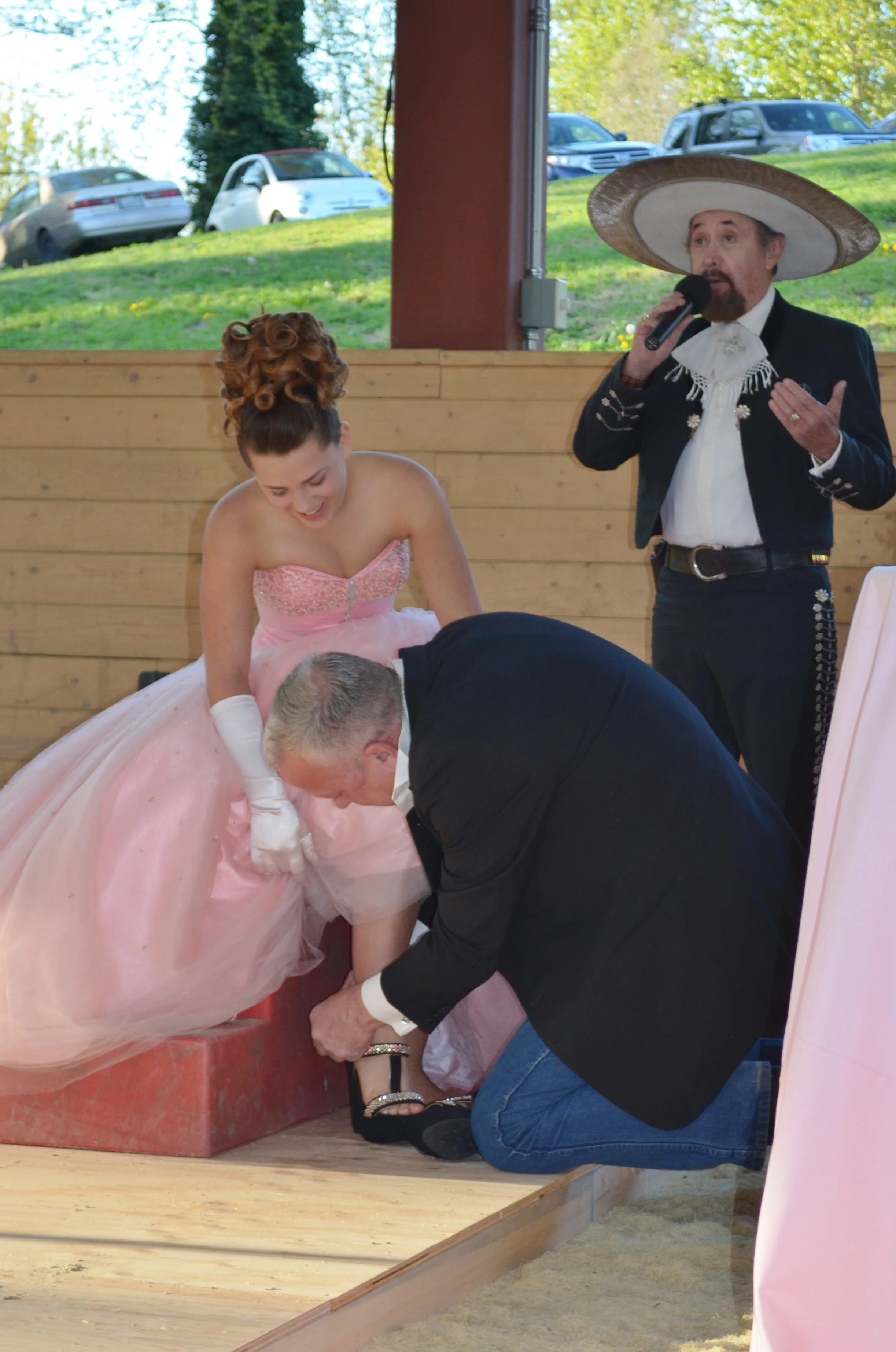A man and woman on a stage during a wedding ceremony, with a woman dressed in a pink dress and gloves, and a man helping her put on her high heel shoe while a man in a large sombrero speaks into a microphone.