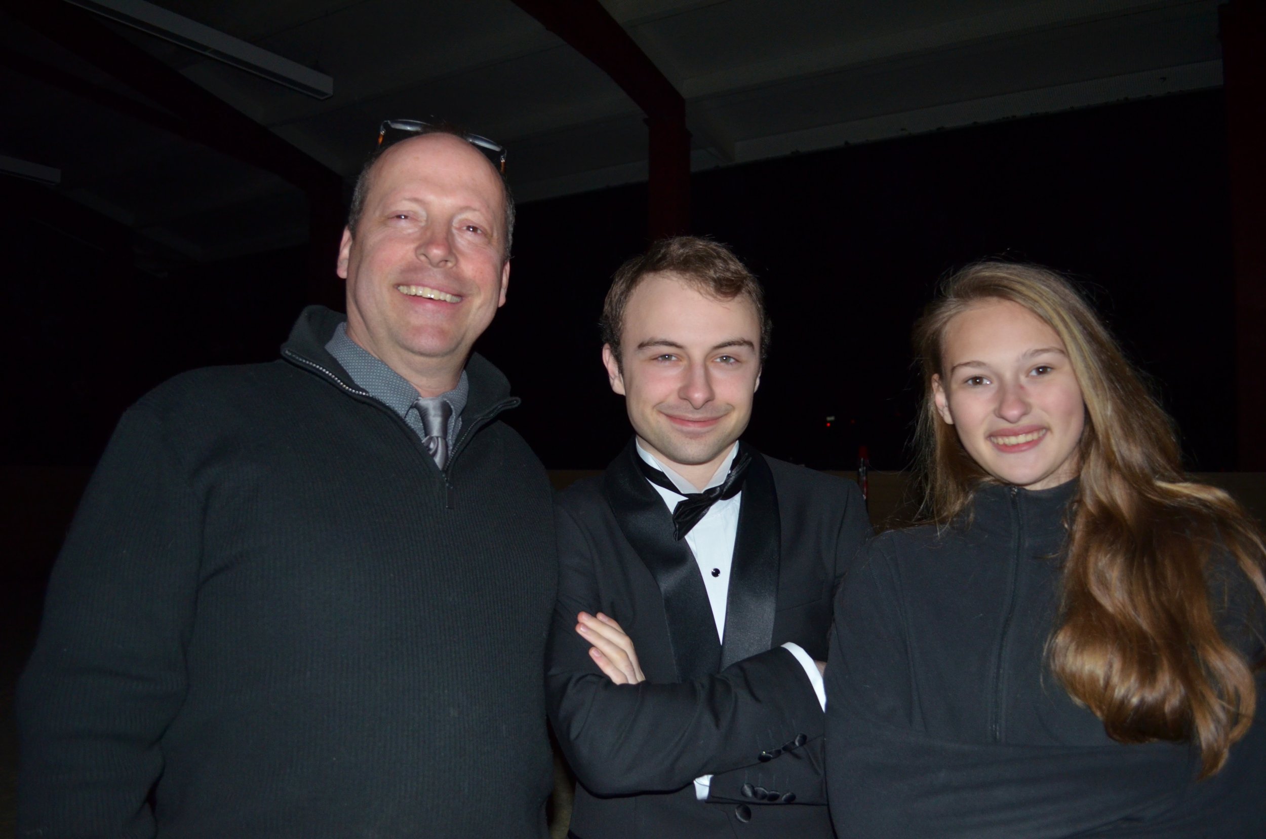Three people smiling at a formal event, with a man on the left wearing a black sweater, a young man in the middle wearing a tuxedo, and a young woman on the right with long hair and a black jacket.