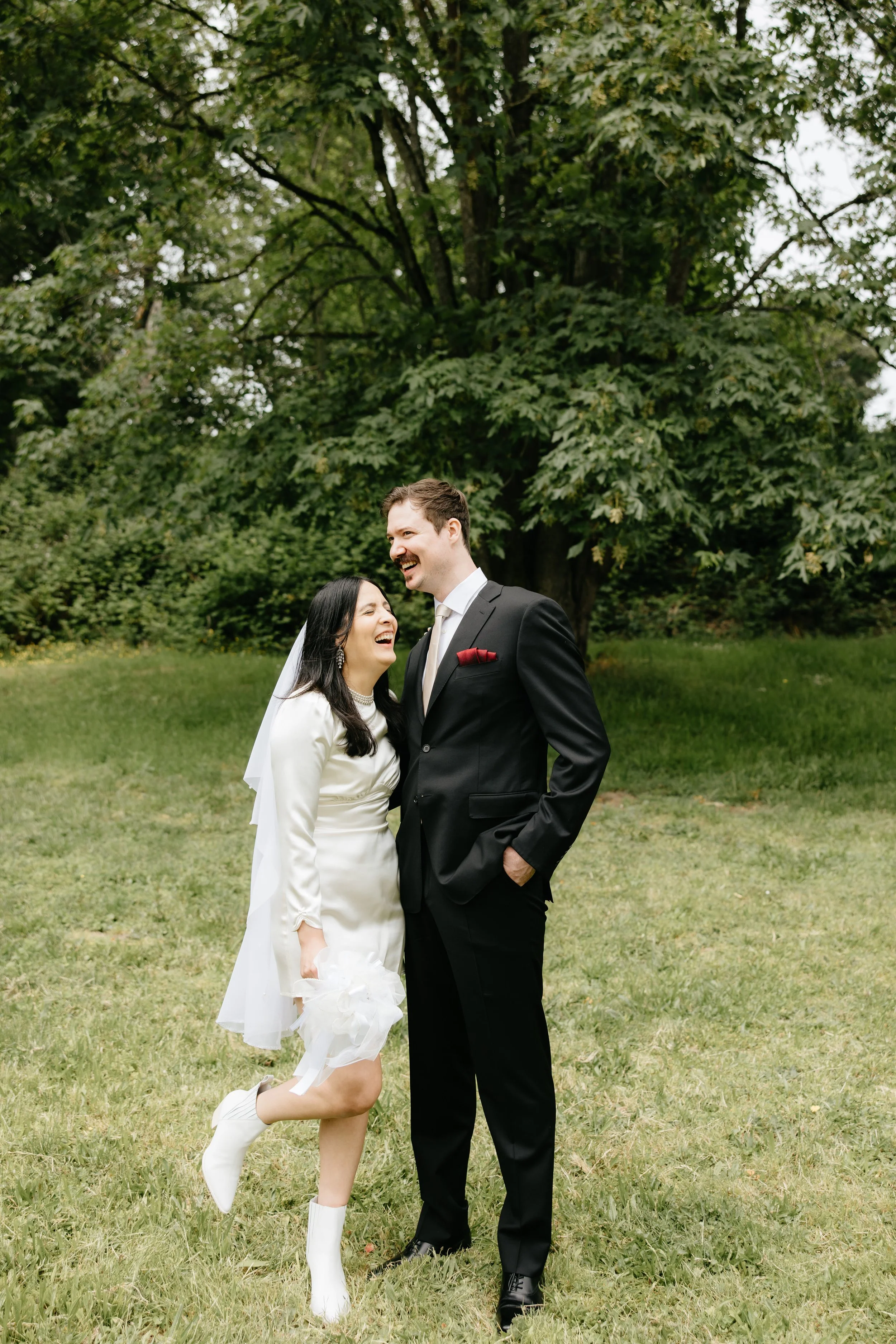 Bride and groom laughing together outdoors on a grassy area with trees in the background.