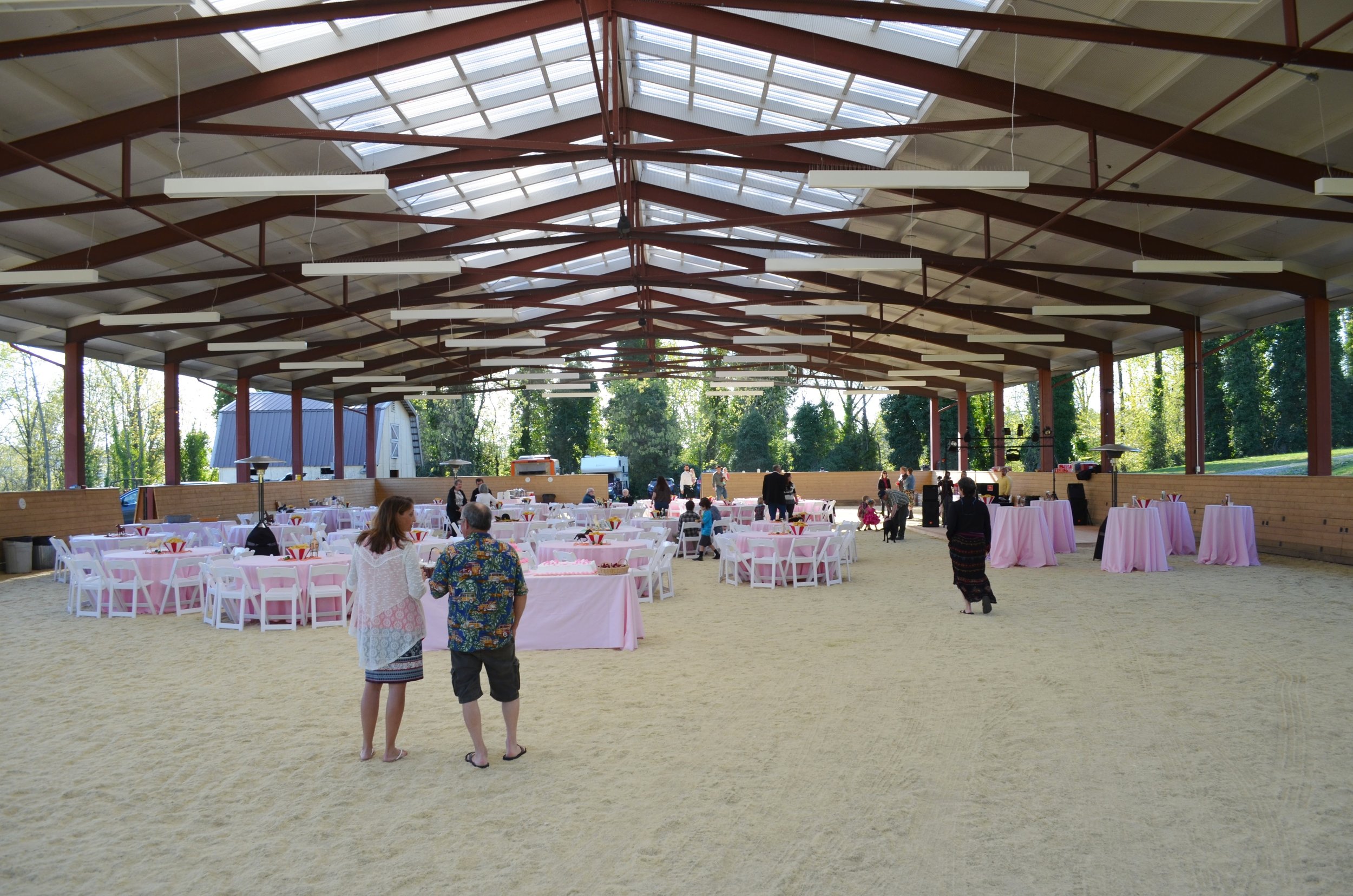 Indoor event space with set tables and chairs, pink tablecloths, and a sandy floor, with people arranging and socializing under a large roof with skylights and wooden beams outside of a green landscape.
