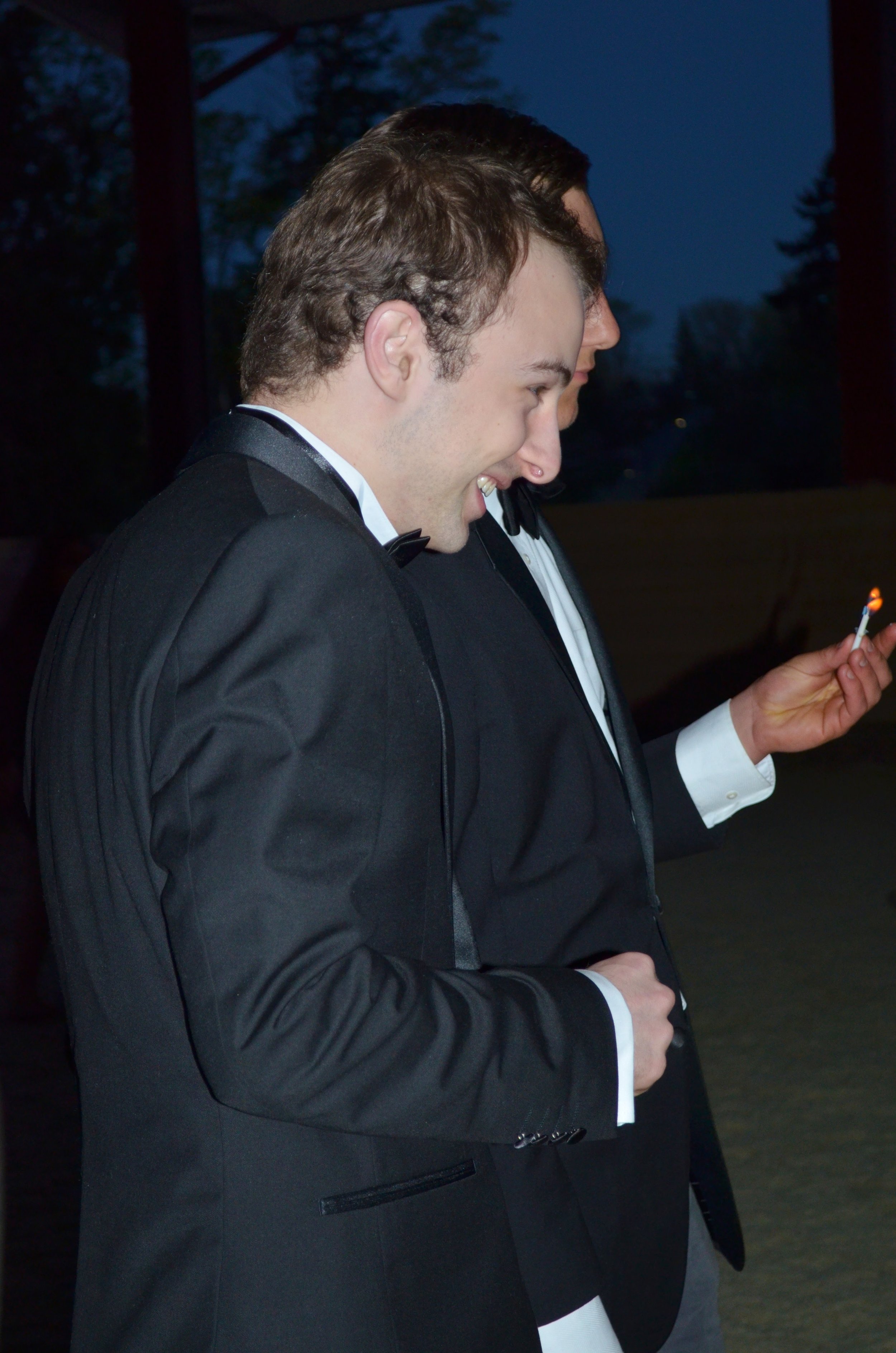 Two men in tuxedos with bow ties smiling and looking at a lit matchstick outdoors during evening or night.