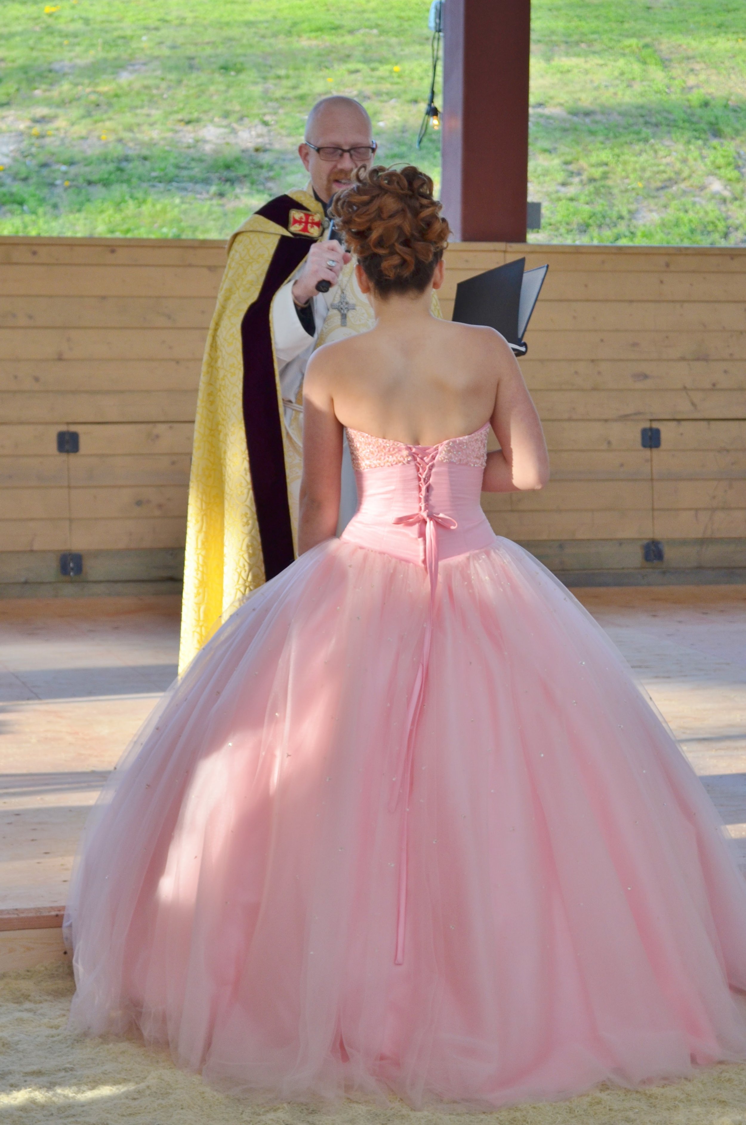 A woman in a pink ball gown getting married in front of a minister in a wooden chapel with greenery outside.