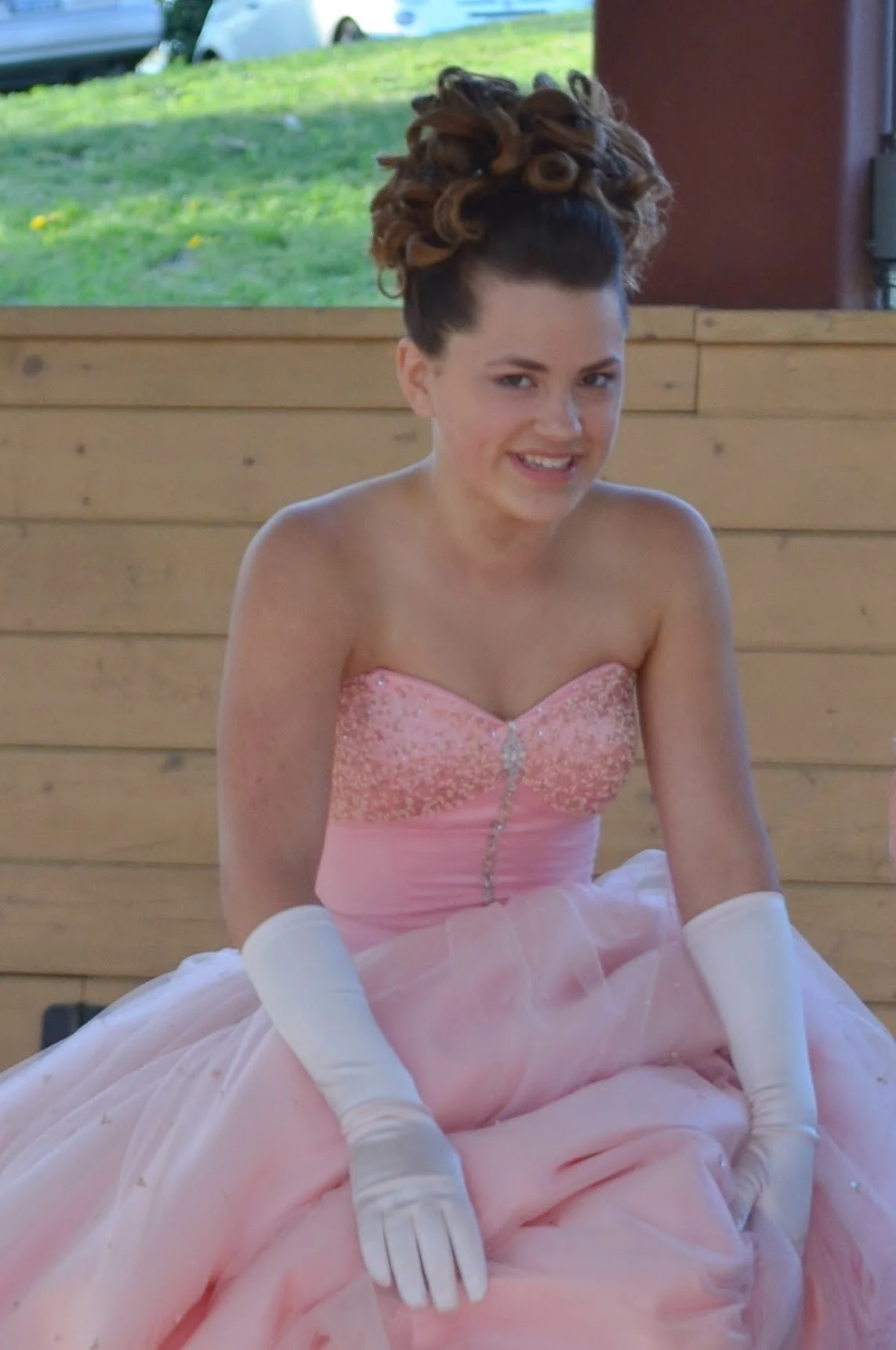 A young woman sitting indoors, wearing a pink strapless ball gown with gloves, with elaborately styled hair.