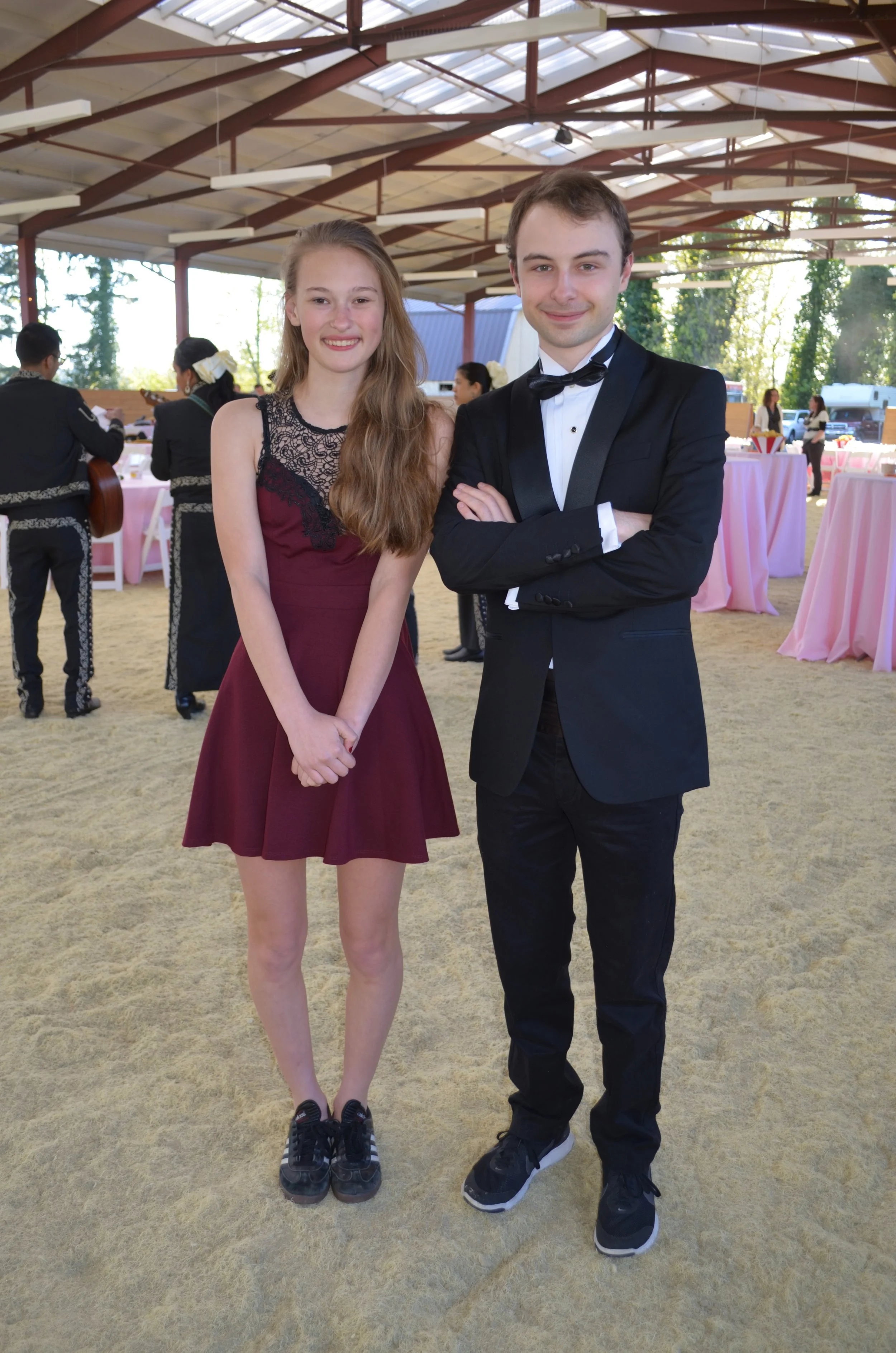 A young woman in a burgundy dress with black lace detail and sneakers, and a young man in a black tuxedo with a bow tie and sneakers, standing together at an indoor event with pink tablecloths and other people in the background.