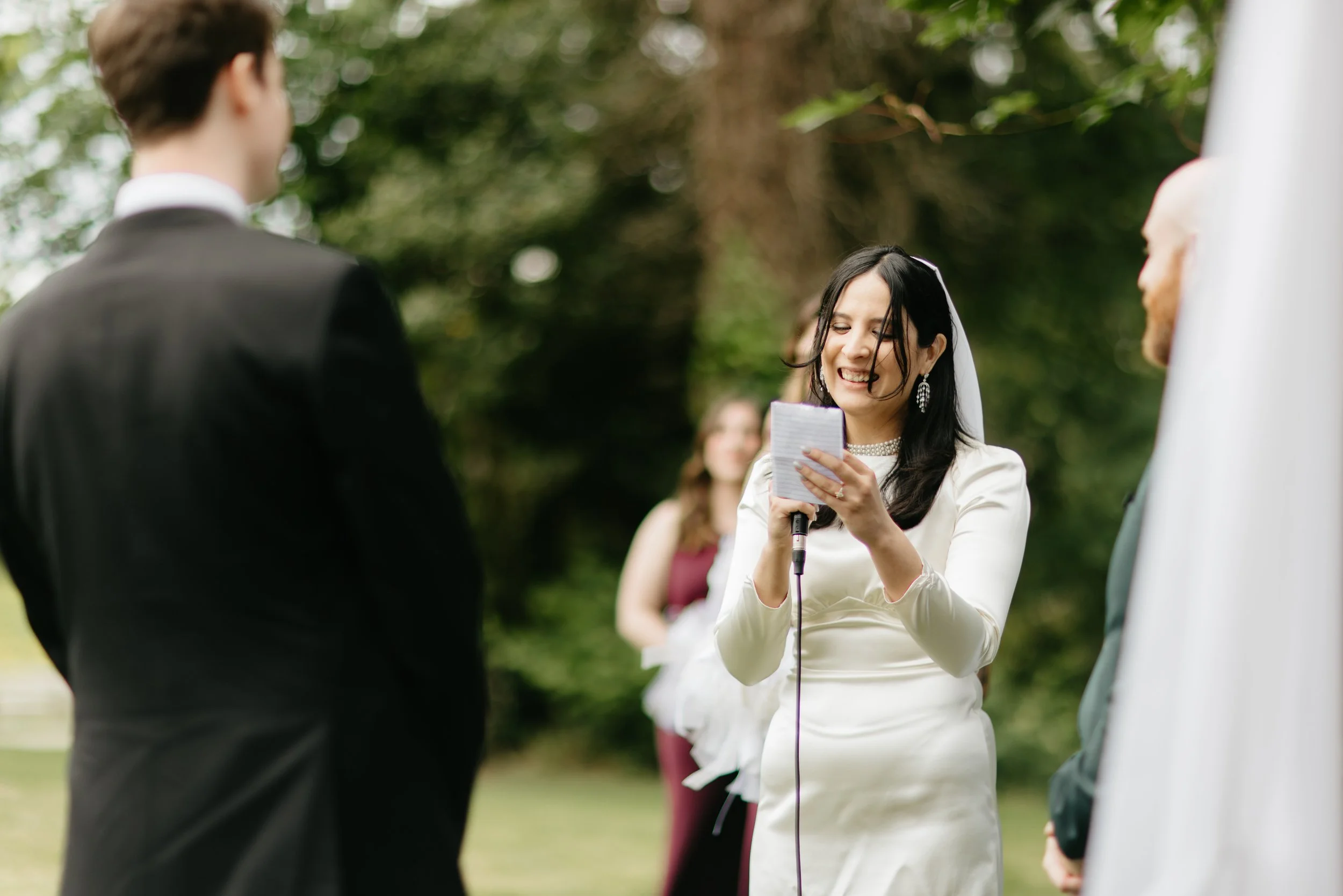 A woman in a white dress reading a vow in a wedding ceremony outdoors, with a bride and groom present.