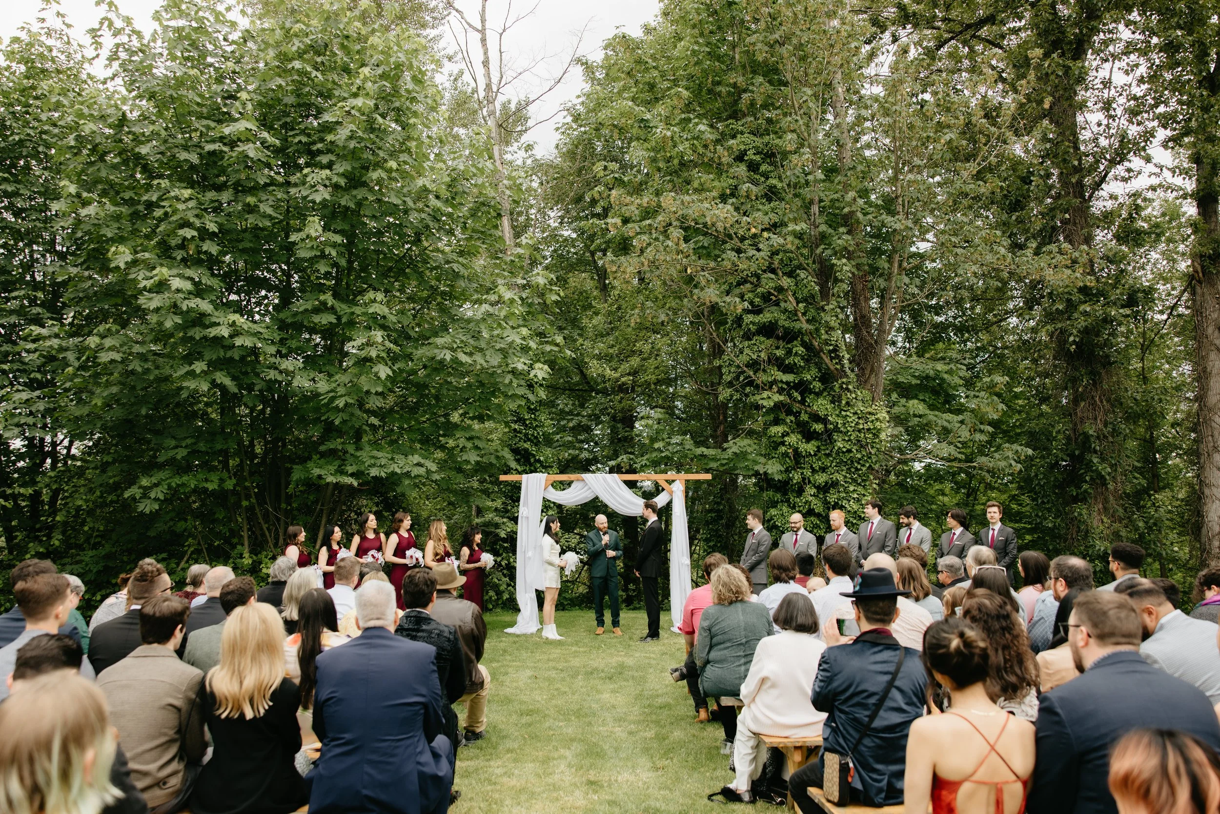 A outdoor wedding ceremony taking place on a grassy area surrounded by tall trees, with guests seated and standing to watch the couple exchange vows under a decorated wooden arch.