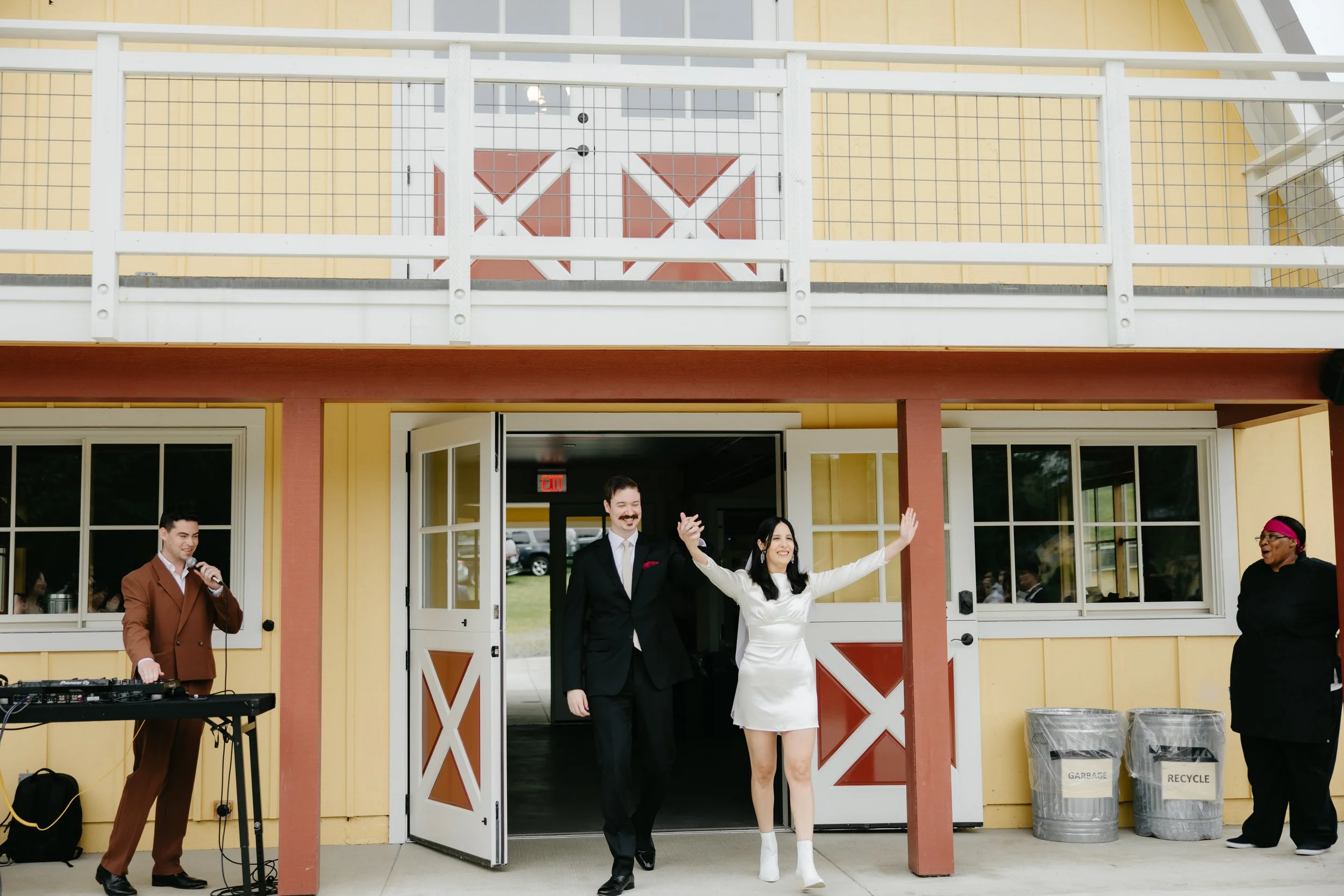 A newlywed couple walking out of a yellow barn-style building with red and white barn doors. The groom is in a black suit, and the bride is in a white dress with white boots, raising her arm in celebration. There is a DJ in a brown suit on the left a