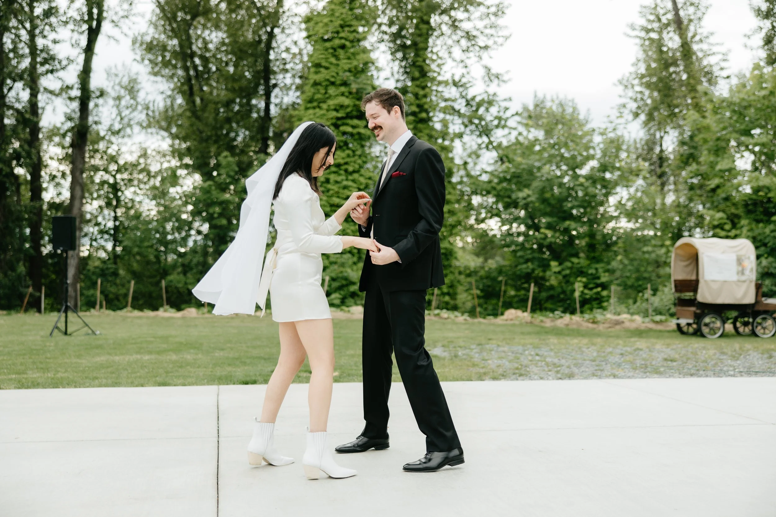 A couple getting engaged outdoors on a paved area, with green trees in the background. The woman is dressed in white with a short dress, white boots, and a veil, while the man is wearing a black suit with a red pocket square. The woman is placing a r