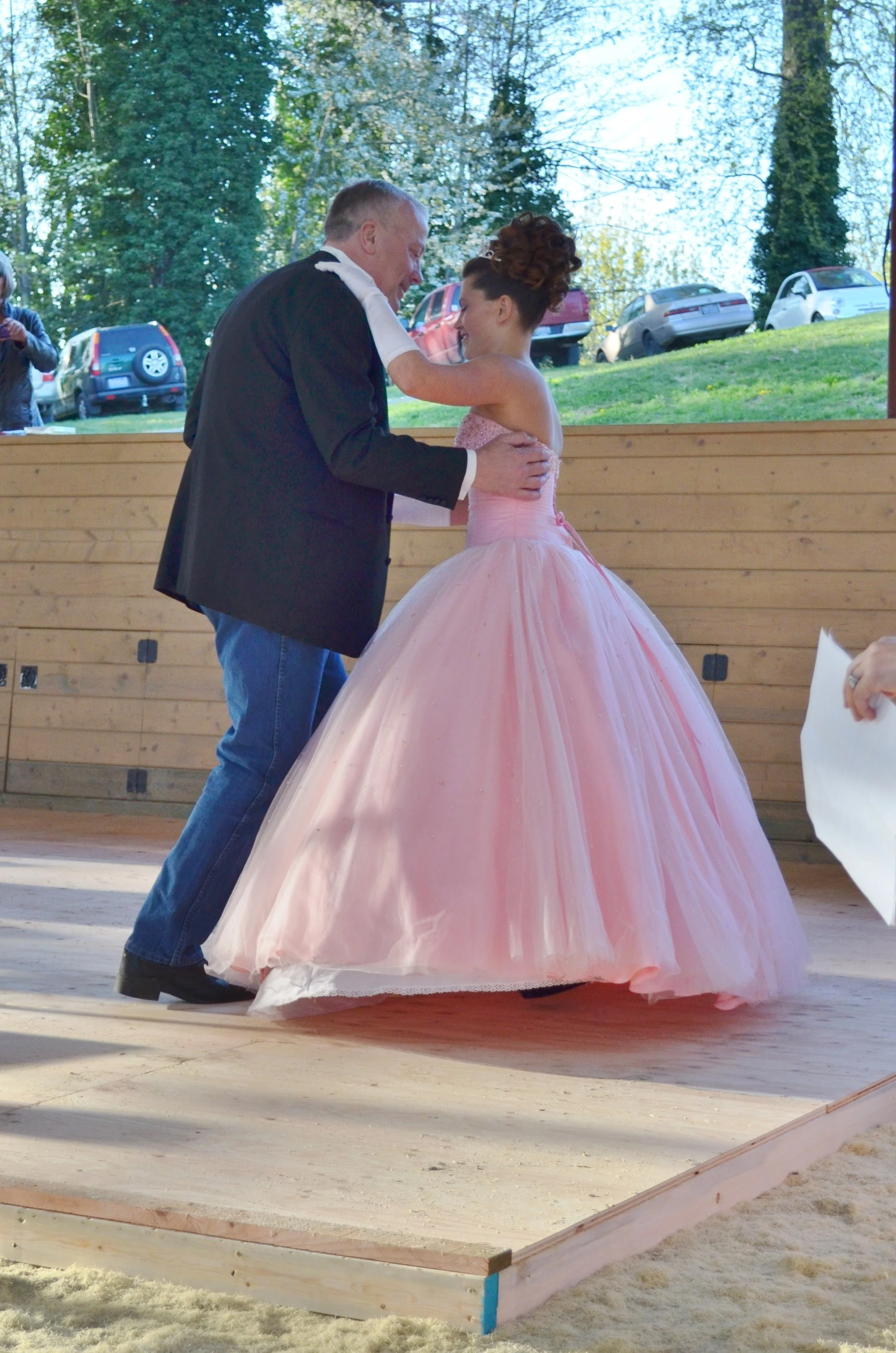 A man and a young girl dance at an outdoor event, the girl wearing a pink ball gown and the man in a black blazer and jeans, with trees and parked cars in the background.