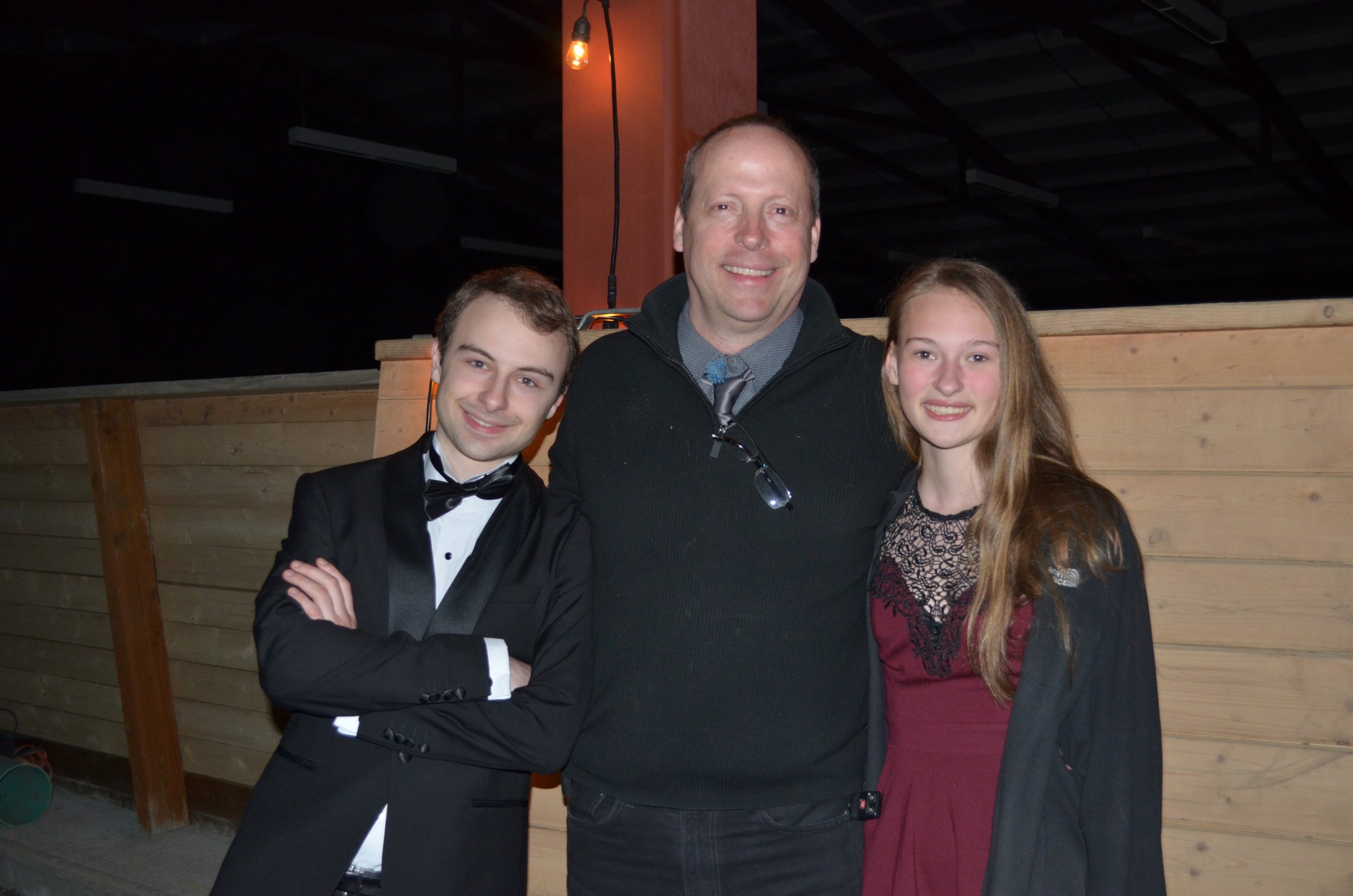 Three people, two young adults and an older man, standing together indoors near a wooden partition, smiling at the camera. The young man on the left wears a tuxedo with a bow tie, the woman on the right wears a burgundy dress with lace detail, and th