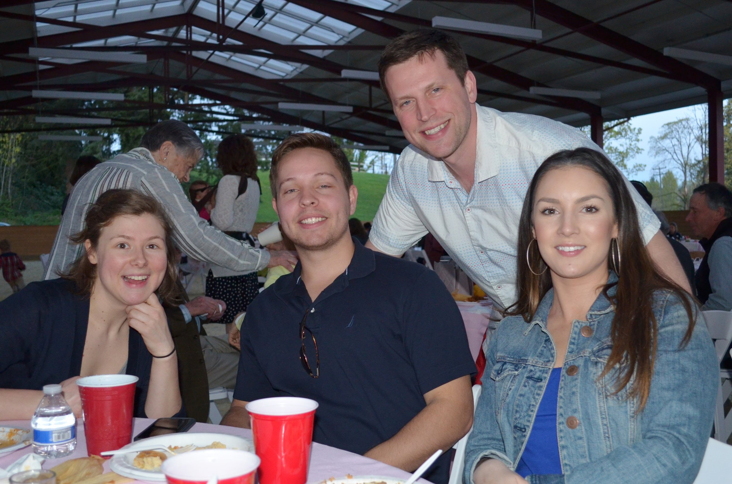 Group of four young adults smiling at an outdoor gathering, with plates of food and drinks on the table, and a pavilion with a metal roof in the background.