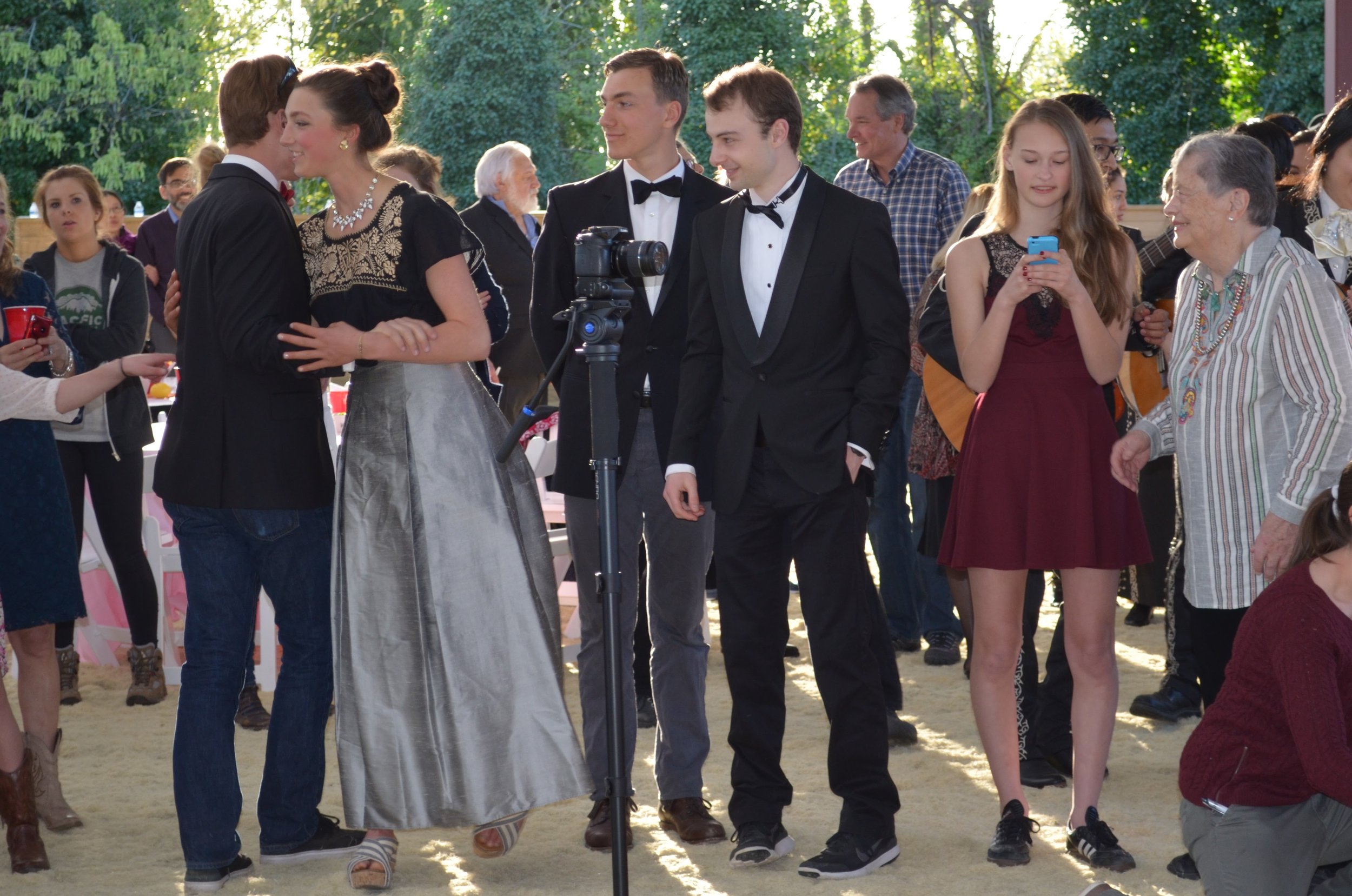 People dressed in formal and casual attire at an outdoor event, with some dancing and others taking photos or using a phone, surrounded by trees and a bright, sunny setting.