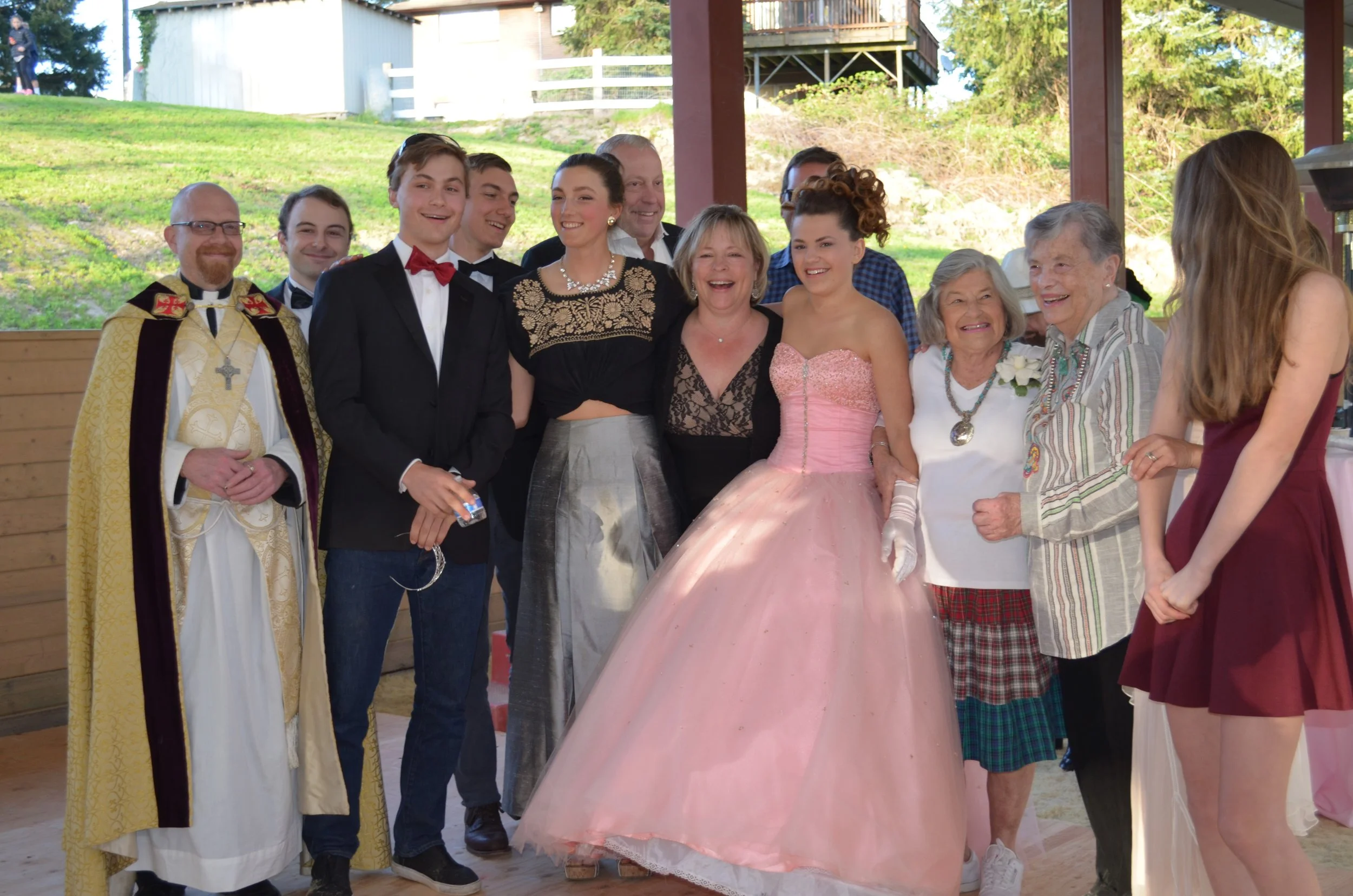 Group of people smiling during a celebration, including a woman in a pink ball gown, and other individuals dressed semi-formally, under a canopy with a grassy outdoor background.