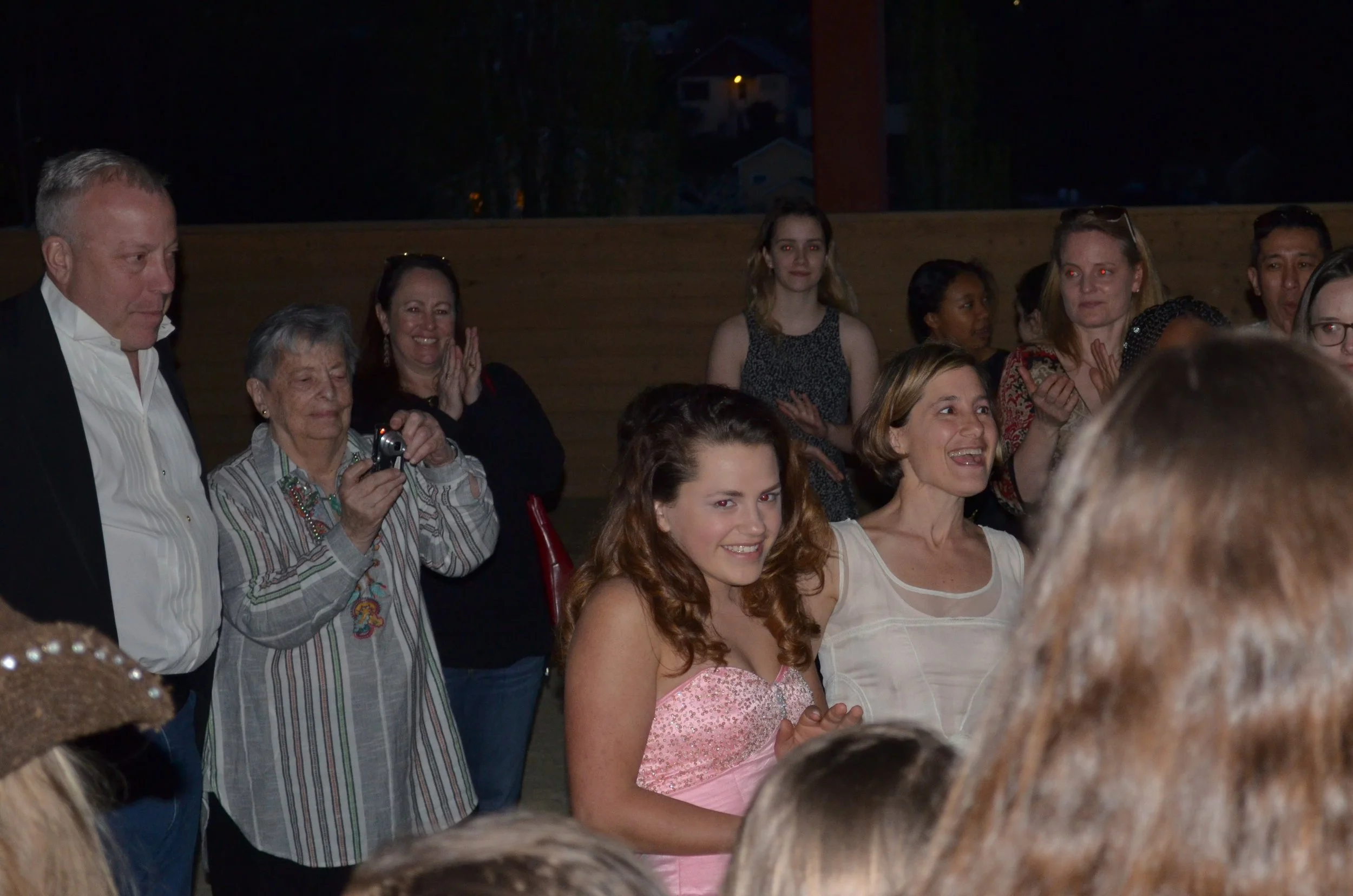 Group of people gathered indoors, some smiling and clapping, with a young woman in a pink dress in the center.