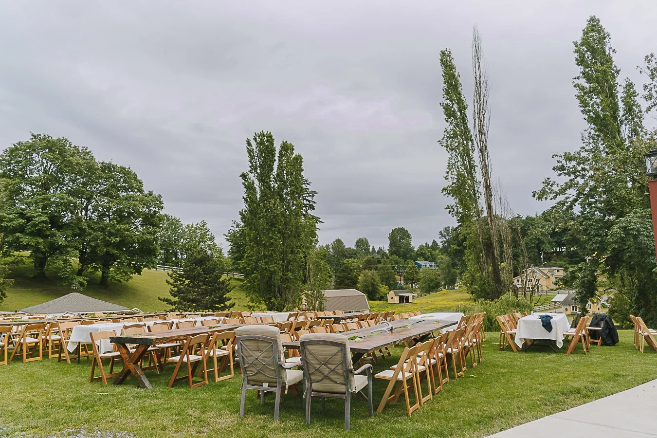 An outdoor event setup with long and small round tables covered with white tablecloths, surrounded by wooden chairs on a grassy lawn. There are tall trees and houses in the background under a cloudy sky.
