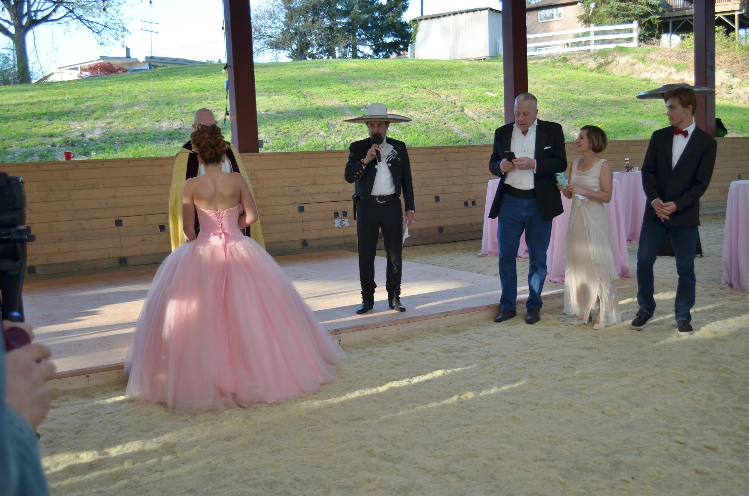 A wedding ceremony taking place outdoors on a sand-covered area. A woman in a pink ball gown stands facing an officiant dressed in a traditional charro suit with a large sombrero. Four other people, including a man in a white shirt with black blazer,