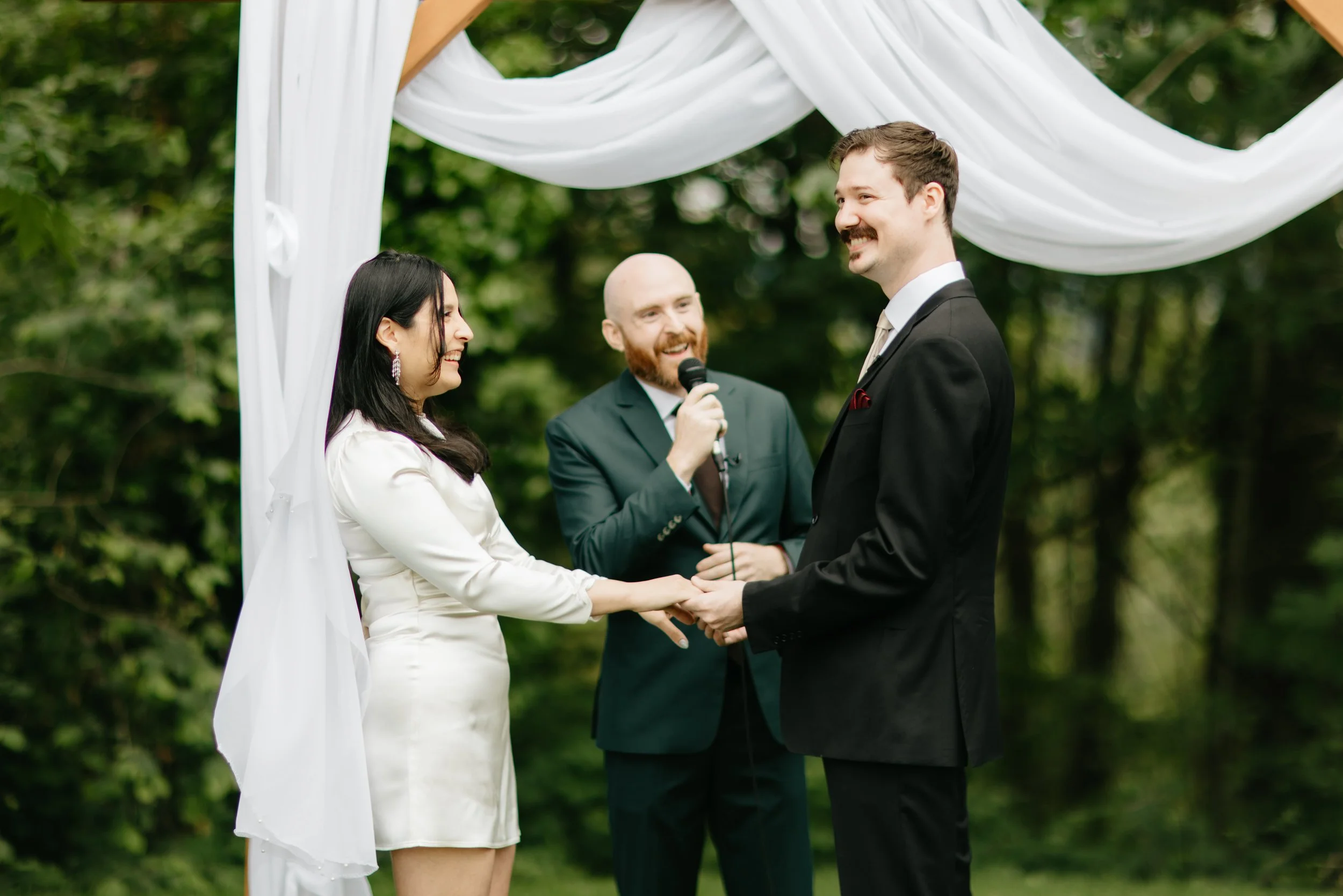 A couple getting married outdoors, holding hands and smiling at each other under a white fabric wedding arch, with a officiant standing behind them holding a microphone, in a green forest setting.