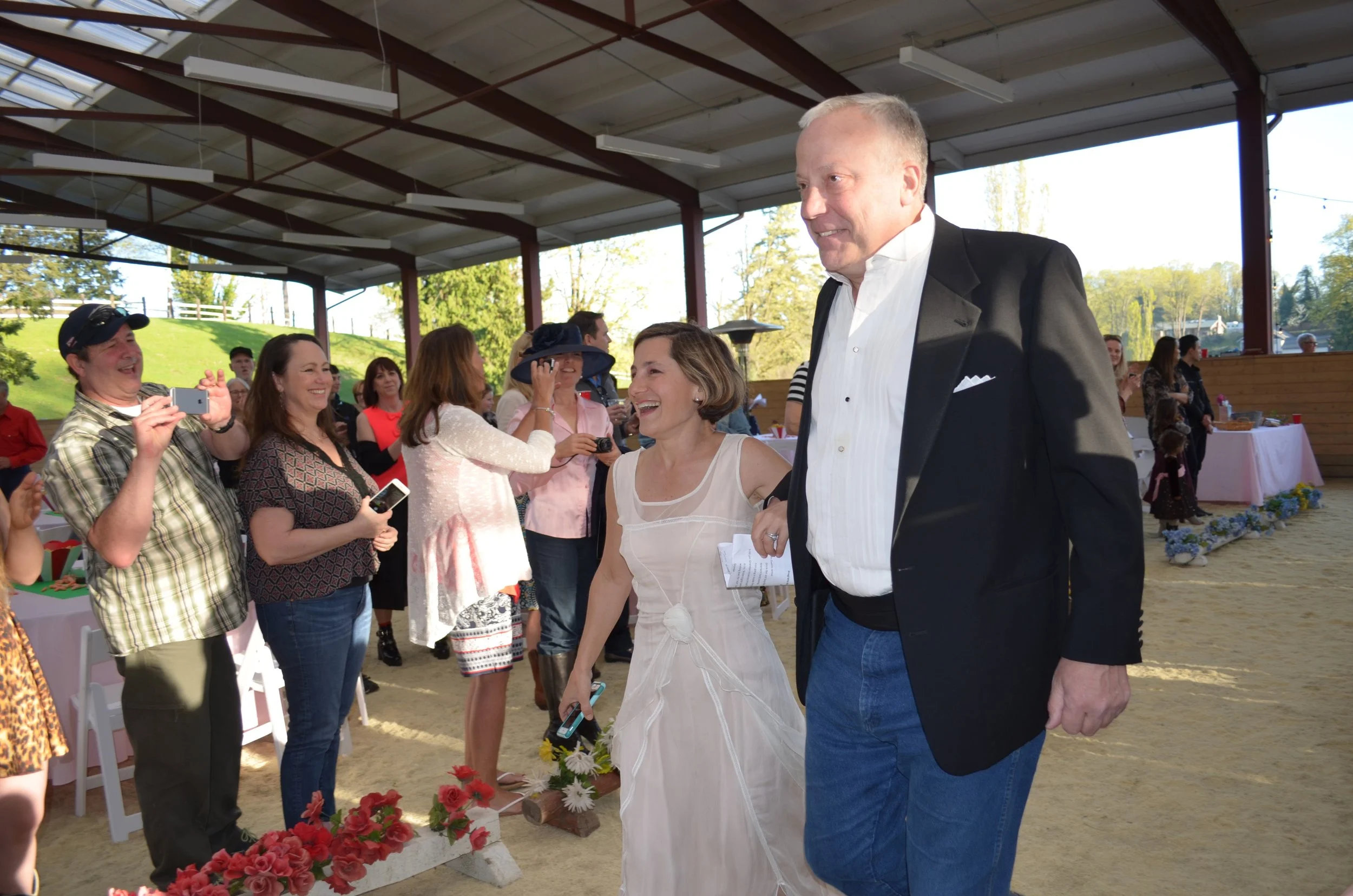 A joyous wedding celebration inside a pavilion with a dirt floor, where a bride in a white dress is holding hands with a tall man in a black tuxedo jacket and blue jeans, smiling as they walk past onlookers taking photos and capturing the moment.