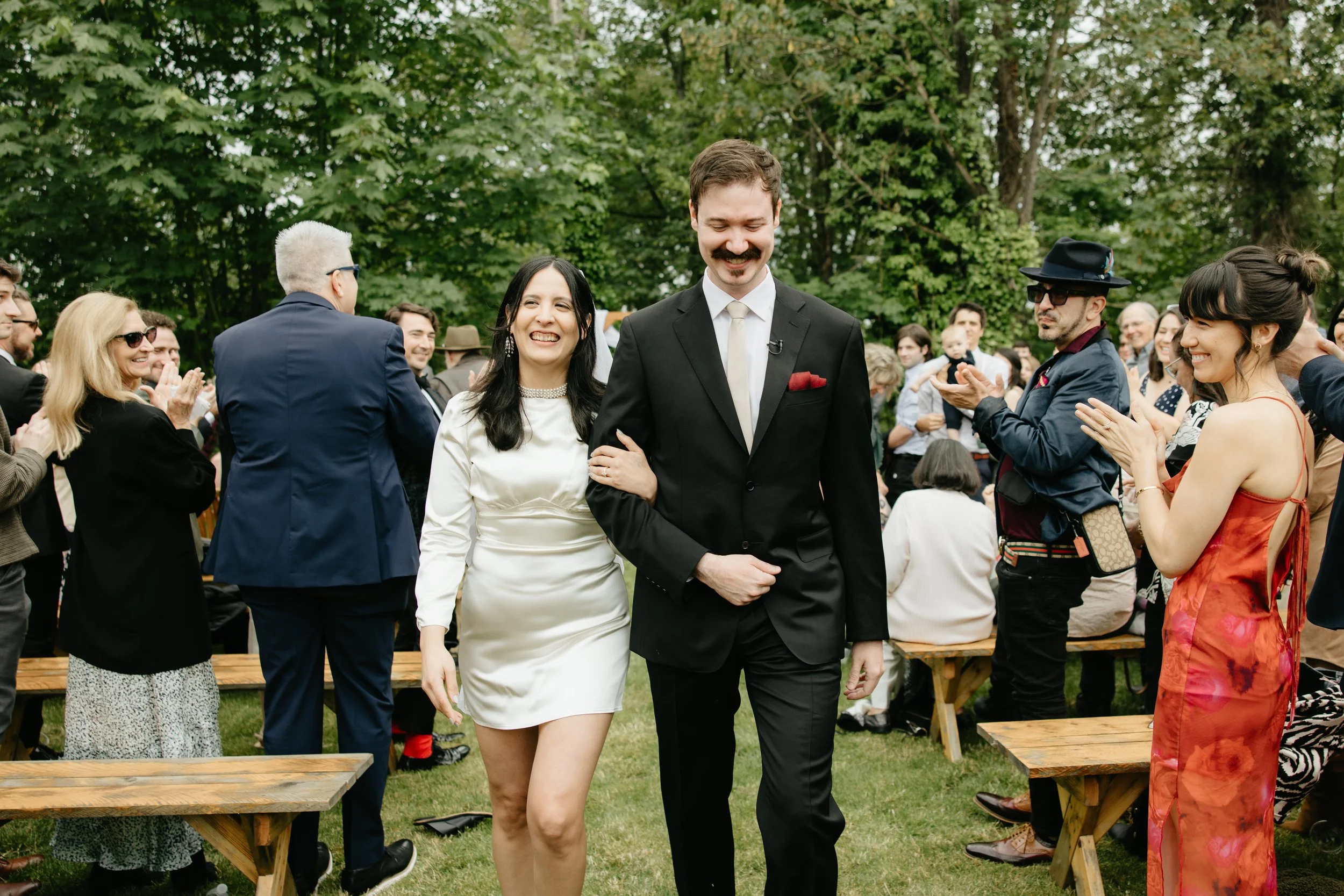 A man in a black suit and a woman in a white dress walking arm-in-arm through an outdoor gathering, with people clapping and smiling around them.