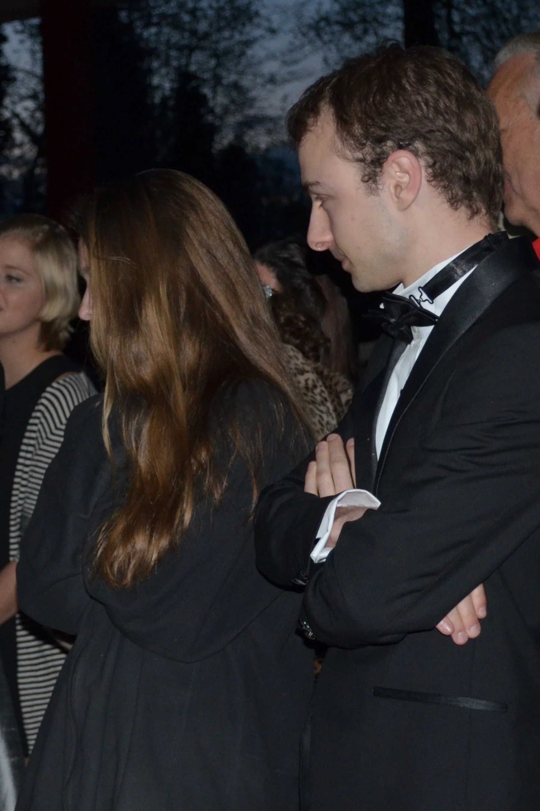 People attending an outdoor evening event, dressed formally, with a man in a tuxedo and bow tie standing with arms crossed.