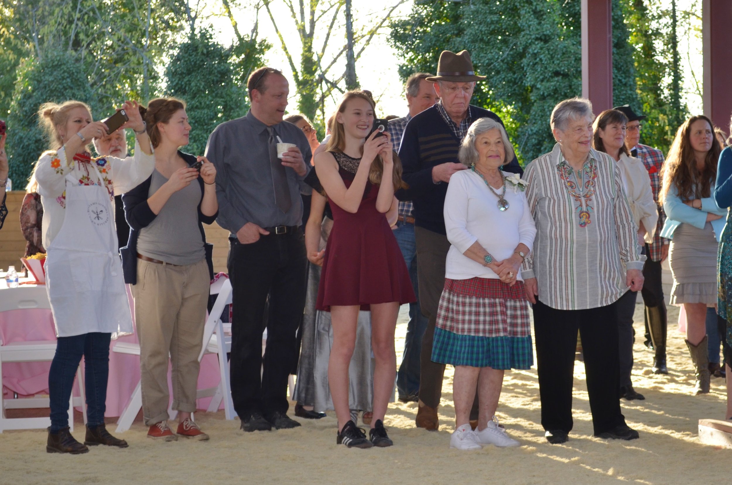 A group of people, including young and elderly individuals, standing outdoors during daytime, some taking photos, with trees and natural scenery in the background.