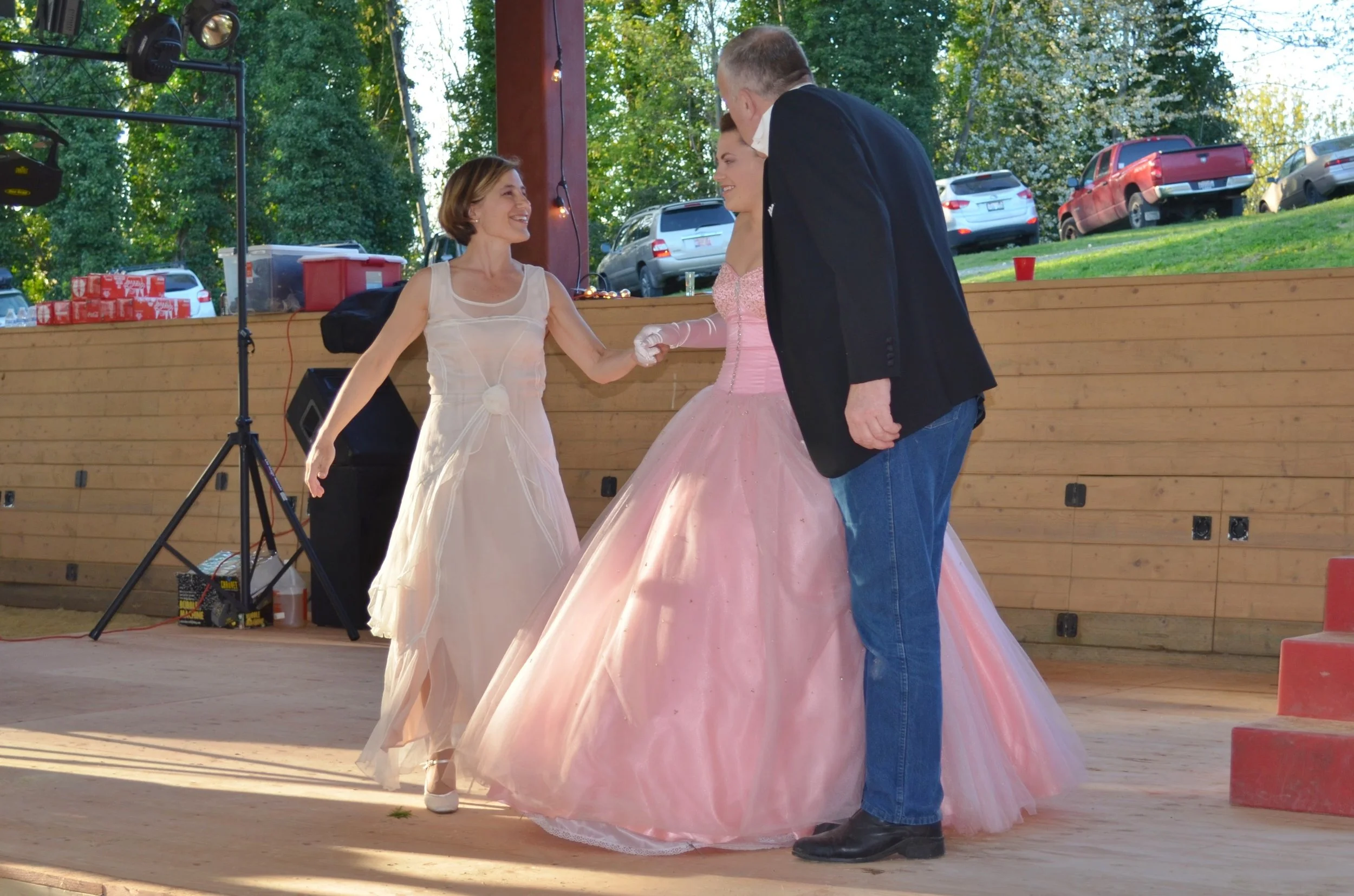 A group of three people dancing at an outdoor event, with a woman in a pink ball gown, a woman in a beige dress, and a man in a dark blazer and jeans. They are smiling and holding hands, with cars and trees in the background.