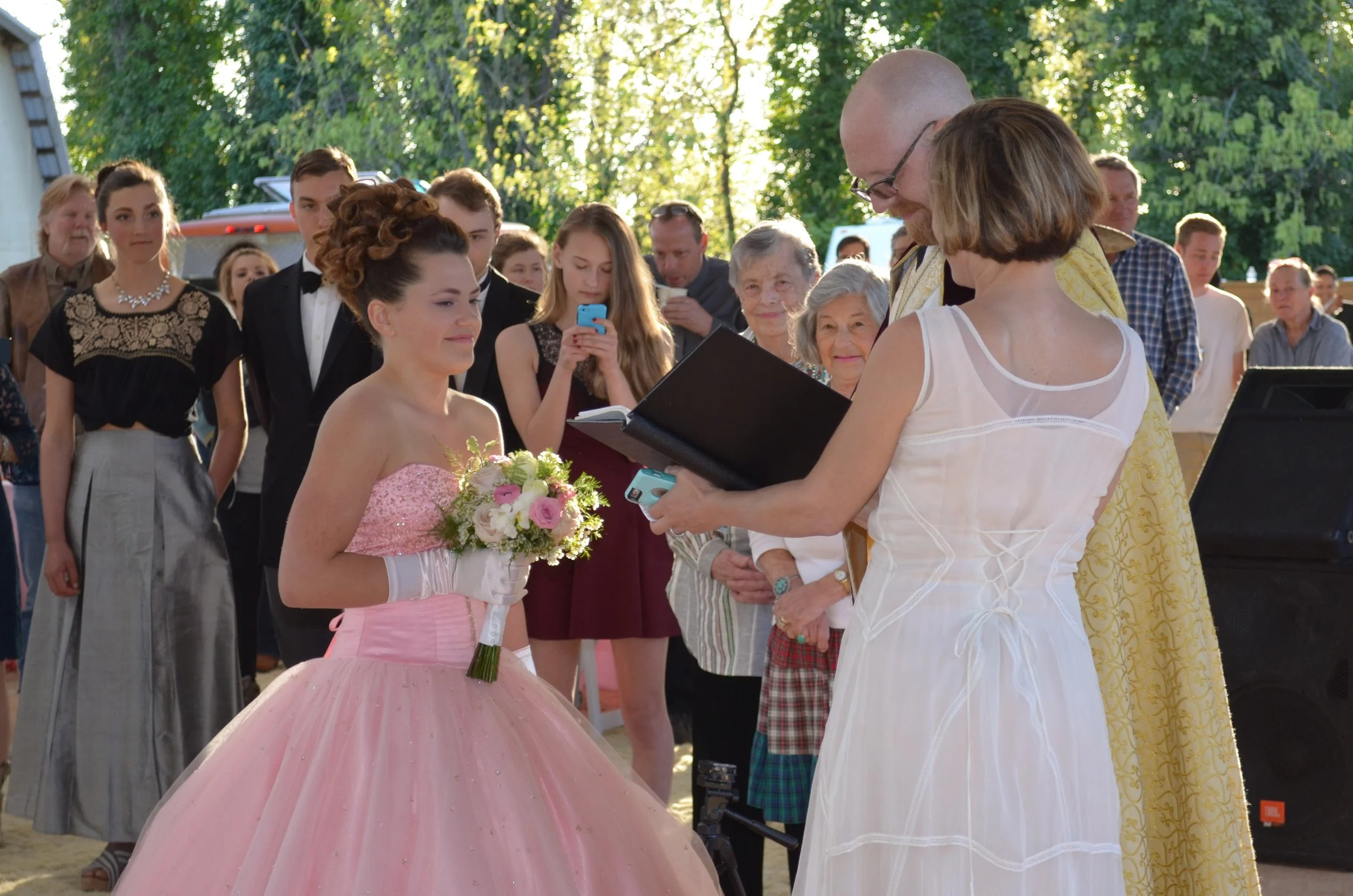 A girl in a pink ball gown receives a bouquet of flowers during a ceremony outdoors, with people watching and taking photos around her.