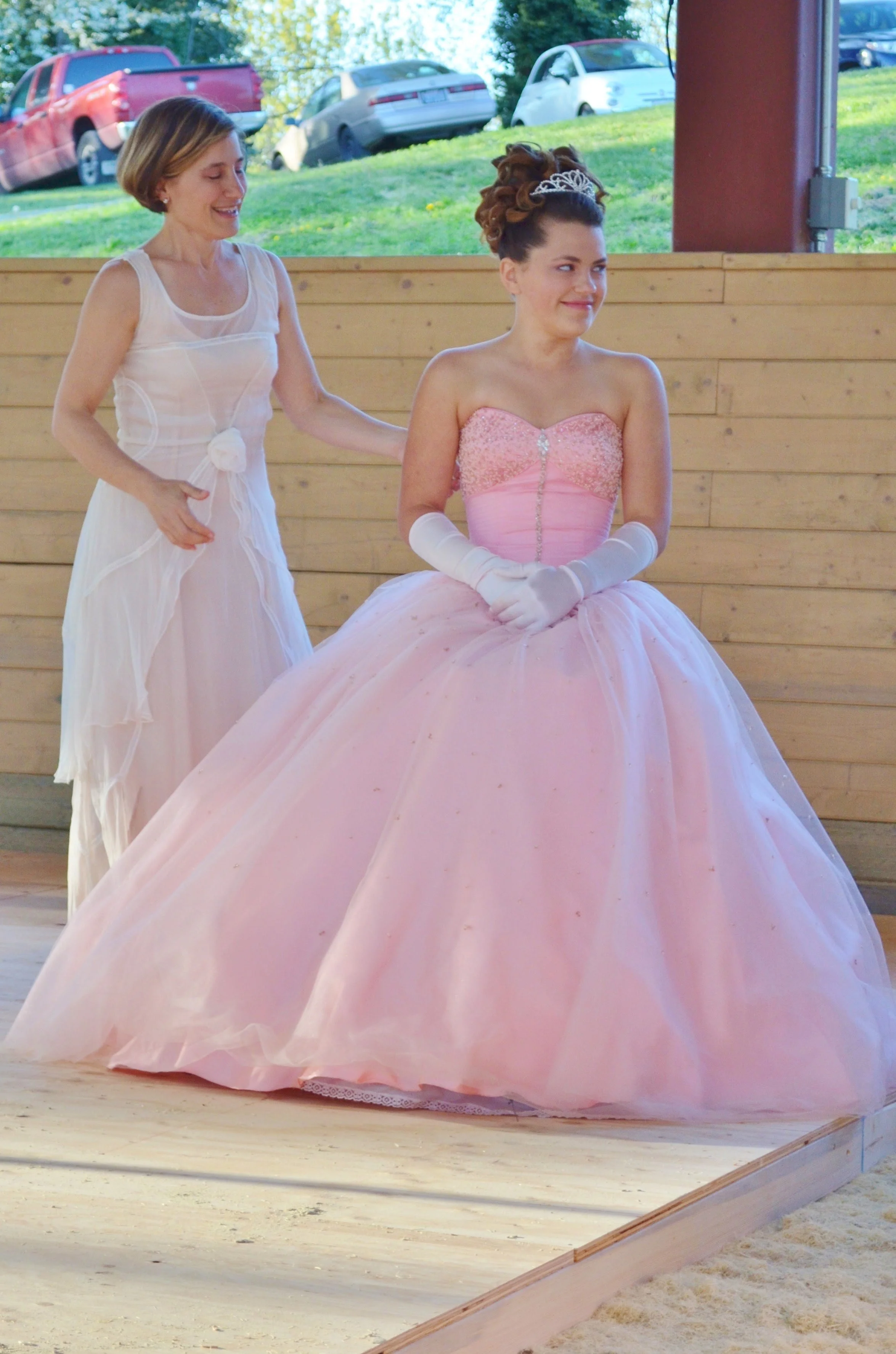 A young woman in a pink ball gown with a tiara and long gloves, standing on a wooden platform, being helped or comforted by an older woman in a white dress, outdoors with parked cars and green grass in the background.