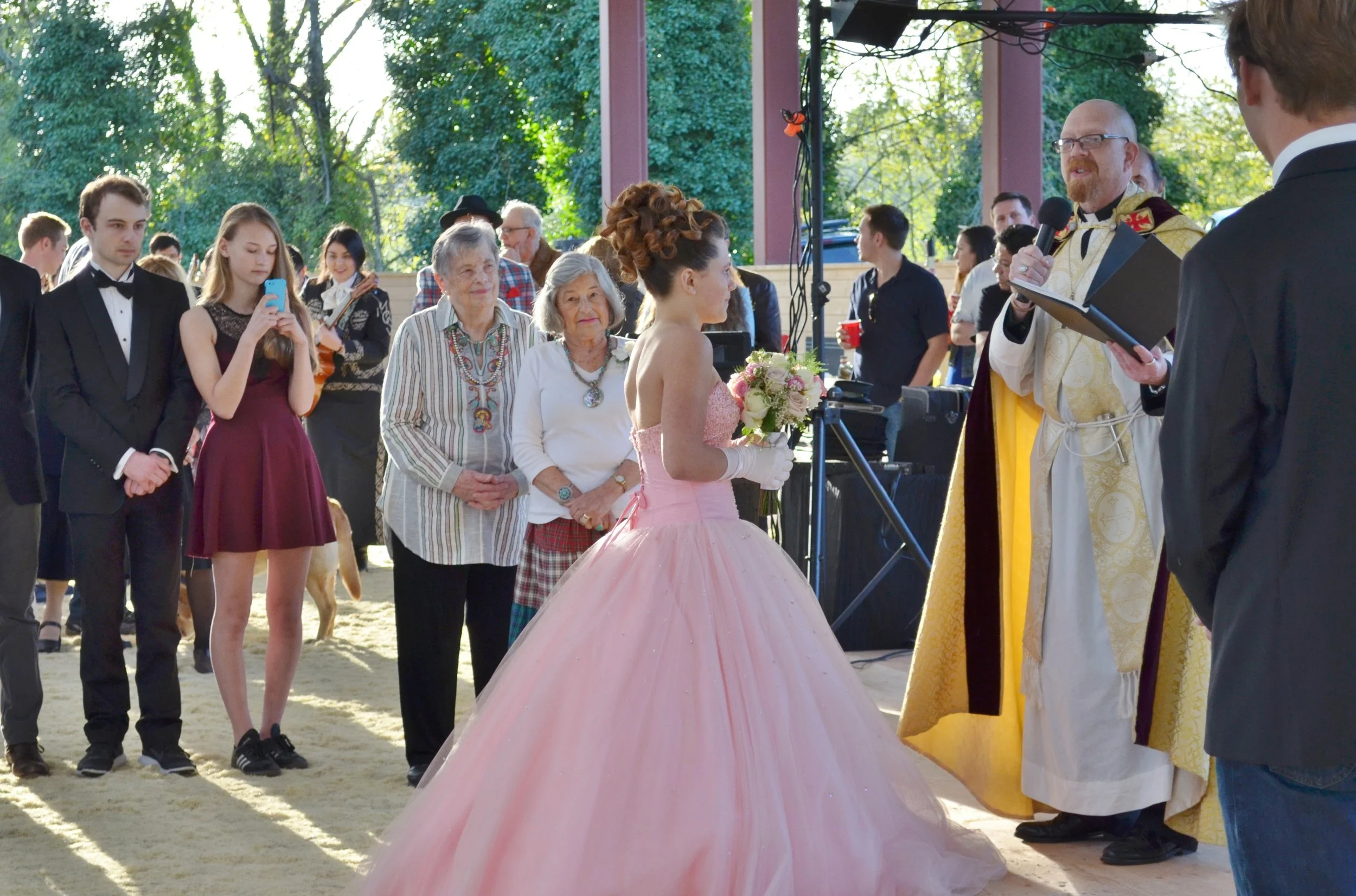 A wedding ceremony with a bride in a pink ball gown holding a bouquet of flowers, standing before a priest in religious robes. The officiant is holding a microphone and reading from a book. Guests, including elderly women and young adults, are watchi